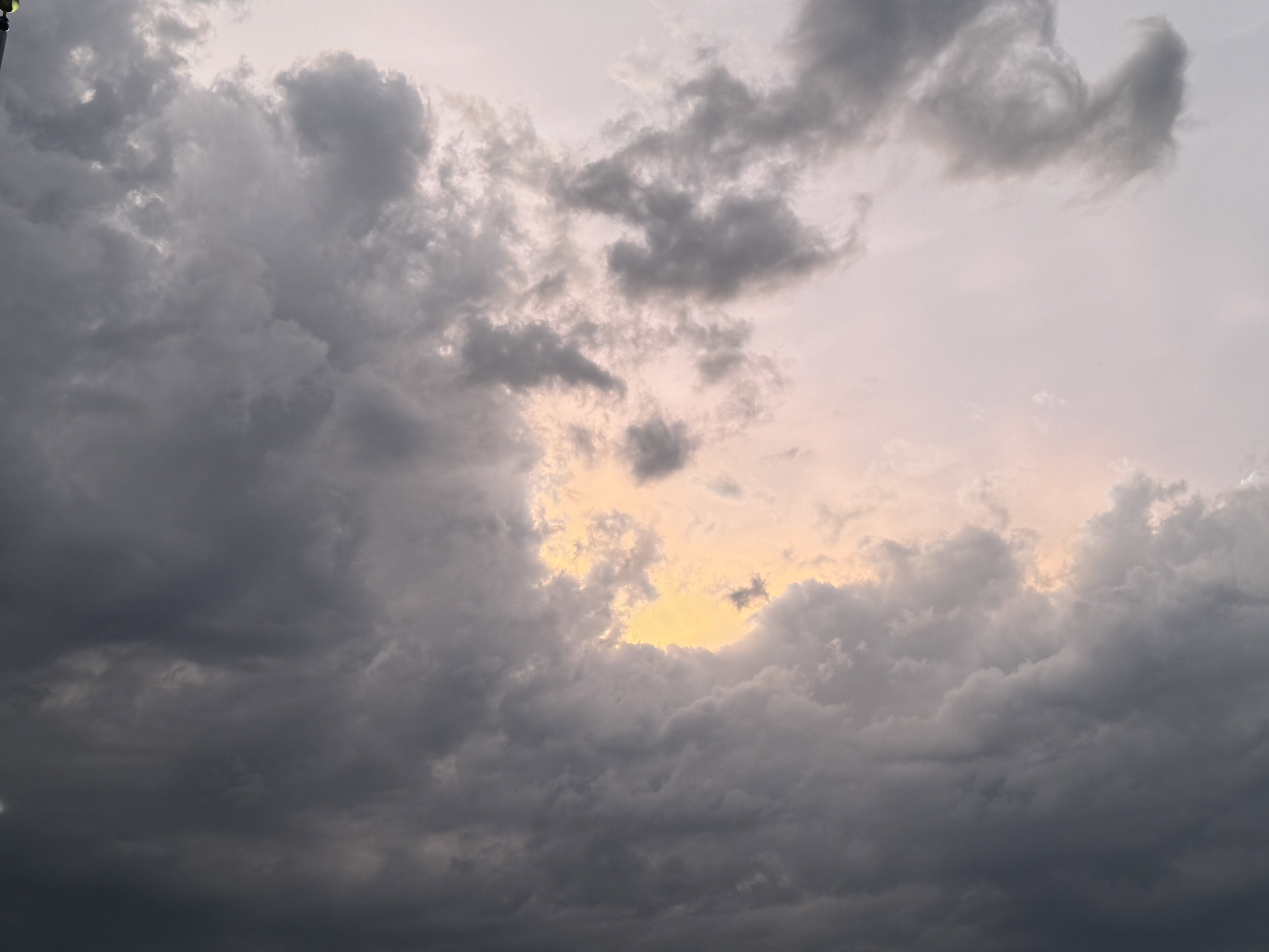 An evening sky framed by thick gray clouds, with a yellow light of the setting sun poking out from the middle, radiating to pink and a purple hue beyond