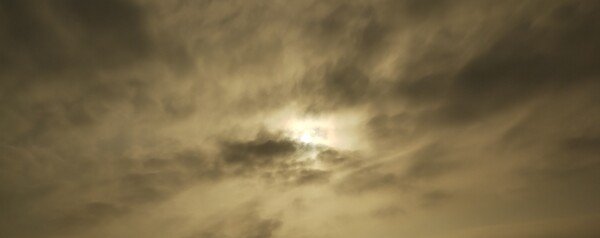 Picture of a kinda beige/gray sky.
The sun's shape is somewhat visible behind a sunlight-covered layer of  darker clouds.