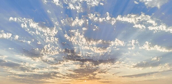a picture of the sunrise with the sun rising from the bottom of the image, with the sky having a gradient from bottom to top colored from orange to a vibrant blue. Clouds are scattered across blocking sun rays slightly creating streaks that allow the sky to be have more vibrant blue colors.