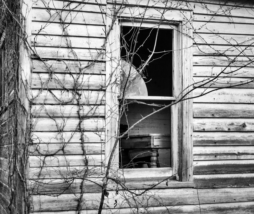 Black and white image of an old, weathered wooden house with peeling paint. A broken window reveals a glimpse of the interior, where a chair is partially visible. Leafless vines creep up the exterior, adding a sense of abandonment and decay.