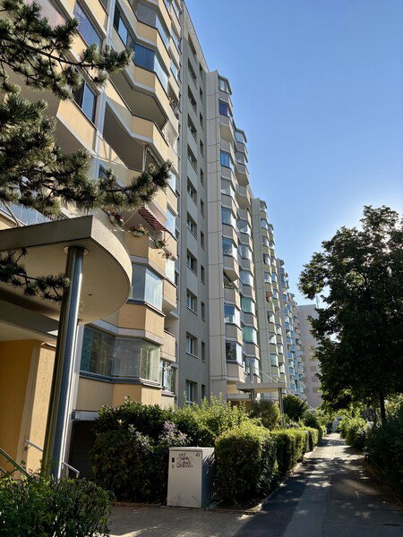 A row of tall, pale residential buildings with many balconies, seen from a low angle on a sunny day. A pathway lined with green bushes runs alongside the buildings.

Eine Reihe von hohen, hellen Wohngebäuden mit vielen Balkonen, von unten an einem sonnigen Tag aufgenommen. Ein von grünen Büschen gesäumter Weg verläuft neben den Gebäuden.