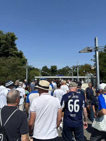 A crowd of soccer fans wearing jerseys and hats walk towards a stadium entrance on a sunny day.

Eine Menge von Fußballfans in Trikots und Hüten geht an einem sonnigen Tag auf den Stadioneingang zu.