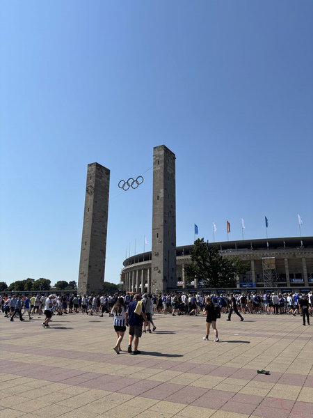 Olympic rings suspended between two large stone towers, with a stadium and a crowd in the background under a clear blue sky.
Olympische Ringe, die zwischen zwei großen Steintürmen hängen, mit einem Stadion und einer Menge im Hintergrund unter einem klaren blauen Himmel.
