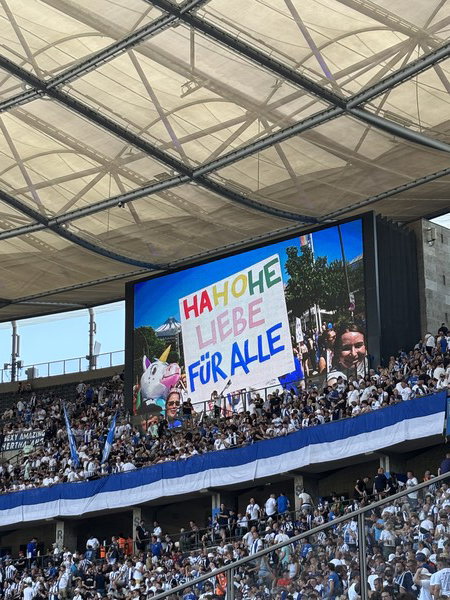 A large screen in a stadium displays a banner with colorful text reading 'HAHOHE LIEBE FÜR ALLE' (High five love for all), accompanied by images of a unicorn and people. A large crowd of spectators wearing white and blue attire is visible in the background, with a blue and white striped banner also displayed.

Ein großer Bildschirm in einem Stadion zeigt ein Banner mit bunten Buchstaben, auf dem 'HAHOHE LIEBE FÜR ALLE' steht, daneben Bilder eines Einhorns und von Menschen. Im Hintergrund ist eine große Menge von Zuschauern in weiß-blauer Kleidung zu sehen, sowie ein blau-weiß gestreiftes Banner.