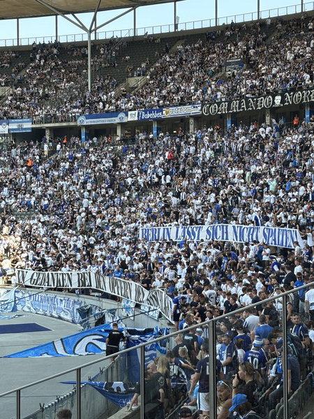 A large crowd of football fans in a stadium, many wearing white and blue, with banners displaying 'COMMANDO NORD' and 'BERLIN LÄSST SICH NICHT SPALTEN'.
Eine große Menge von Fußballfans in einem Stadion, viele in Weiß und Blau gekleidet, mit Bannern, auf denen 'COMMANDO NORD' und 'BERLIN LÄSST SICH NICHT SPALTEN' steht.