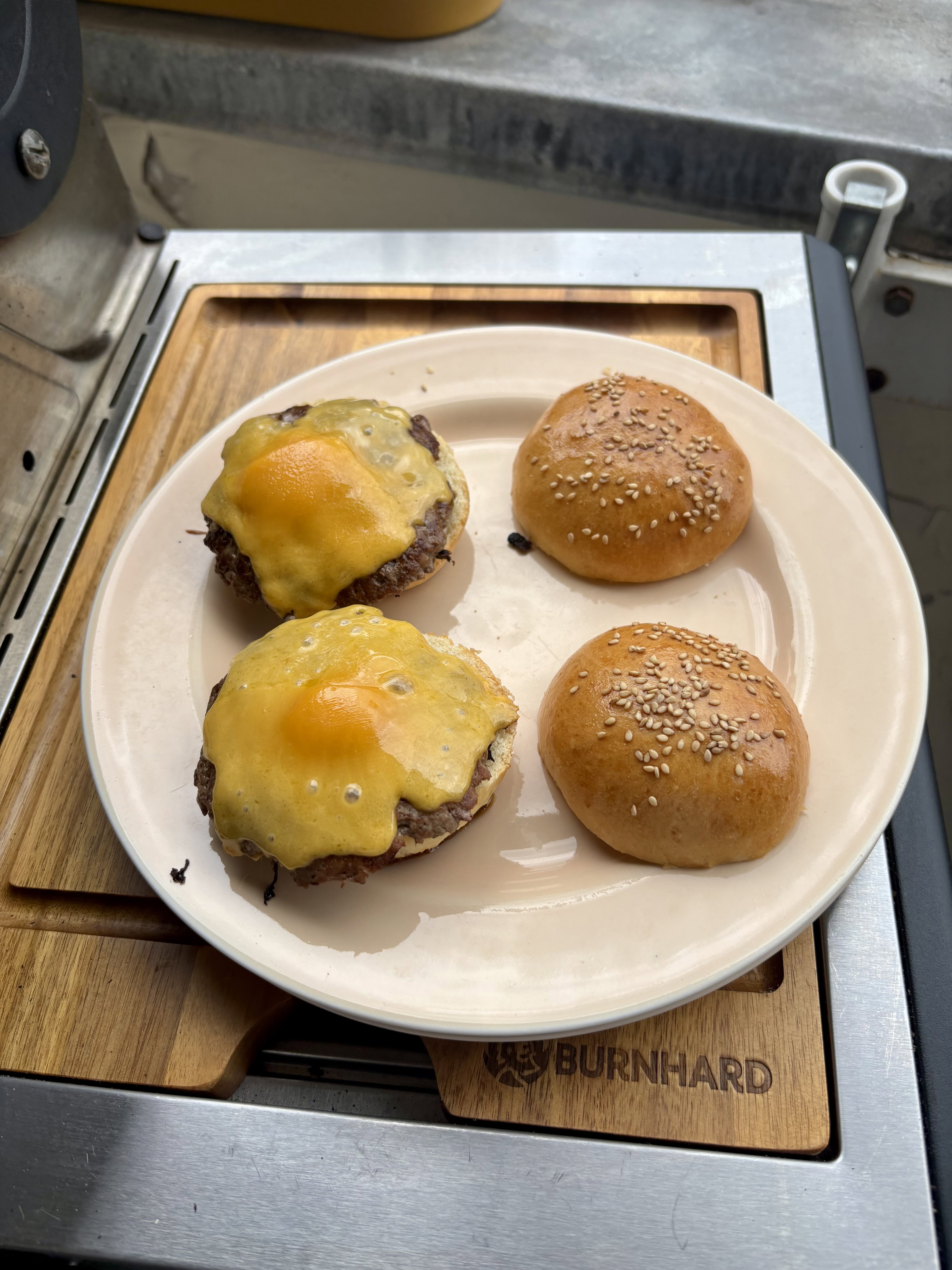 Two cheeseburger patties with melted yellow cheese and two sesame seed burger buns on a plate, resting on a wooden cutting board on a grill. Zwei Cheeseburger-Patties mit geschmolzenem gelben Käse und zwei Brötchen mit Sesam auf einem Teller, auf einem Holzschneidebrett auf einem Grill liegend.