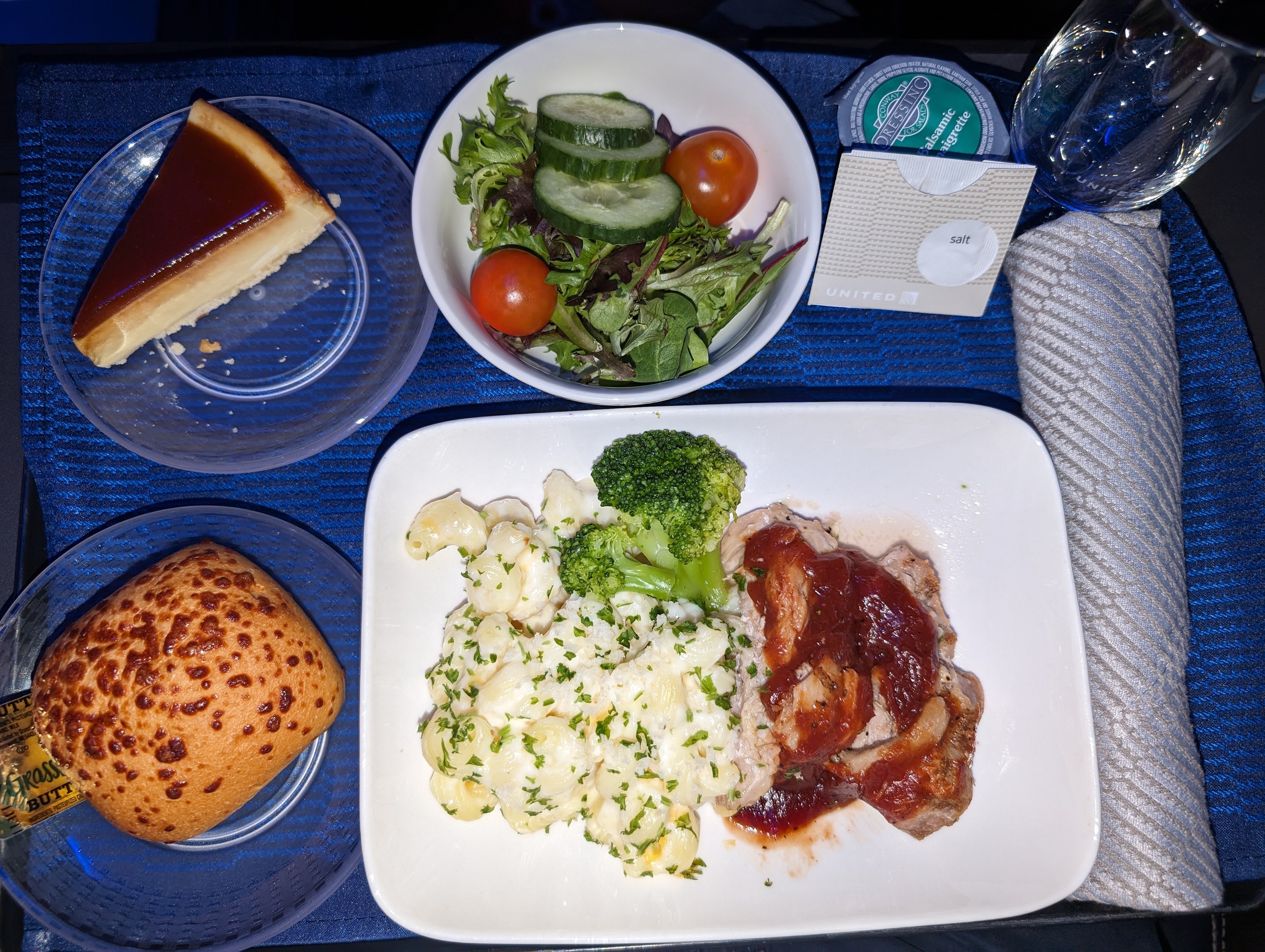 Photo of a tray of food taken on a domestic US United flight. The main entree on the tray is a dish with bbq chicken thighs and macaroni. A salad, roll, and piece of cheesecake are also present.