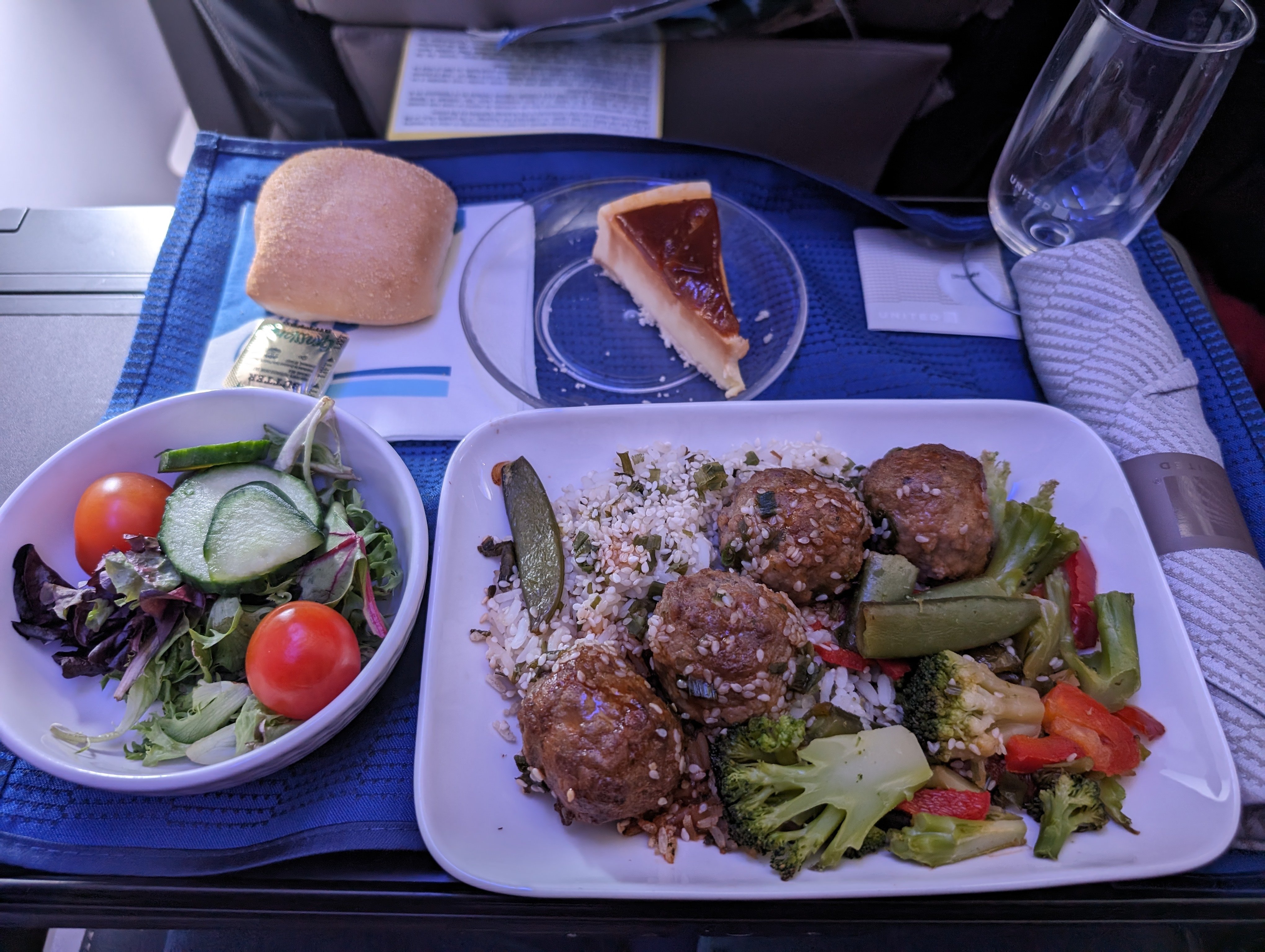 Photo of a tray of food taken on a domestic US United flight. The main entree on the tray is a dish with white rice, plain meatballs, and mixed vegetables. A salad, roll, and piece of cheesecake are also present.