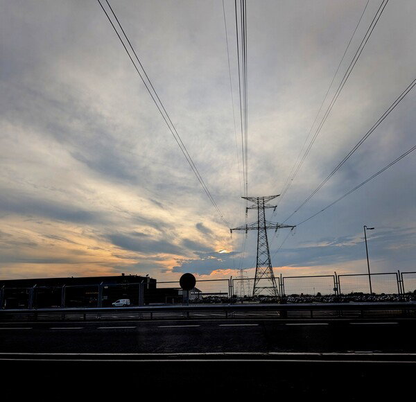 An electrical pylon set against the setting sun. Its wiring leads towards the barely visible shine of the star under a layer of wispy, bluish clouds.
