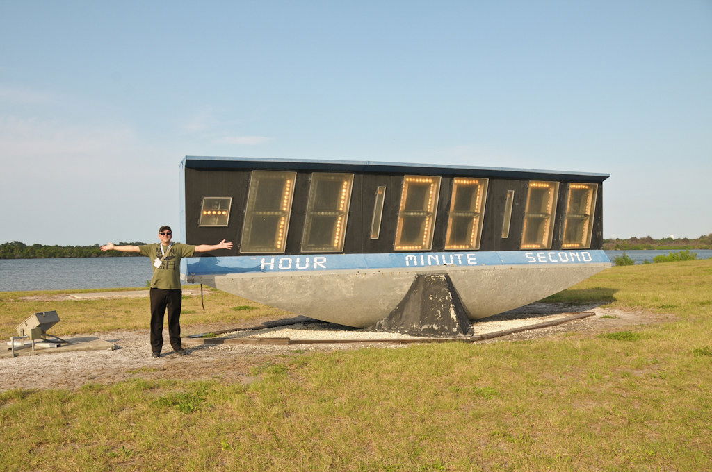 Wide shot of a man standing outdoors with outstretched arms next to an enormous countdown clock