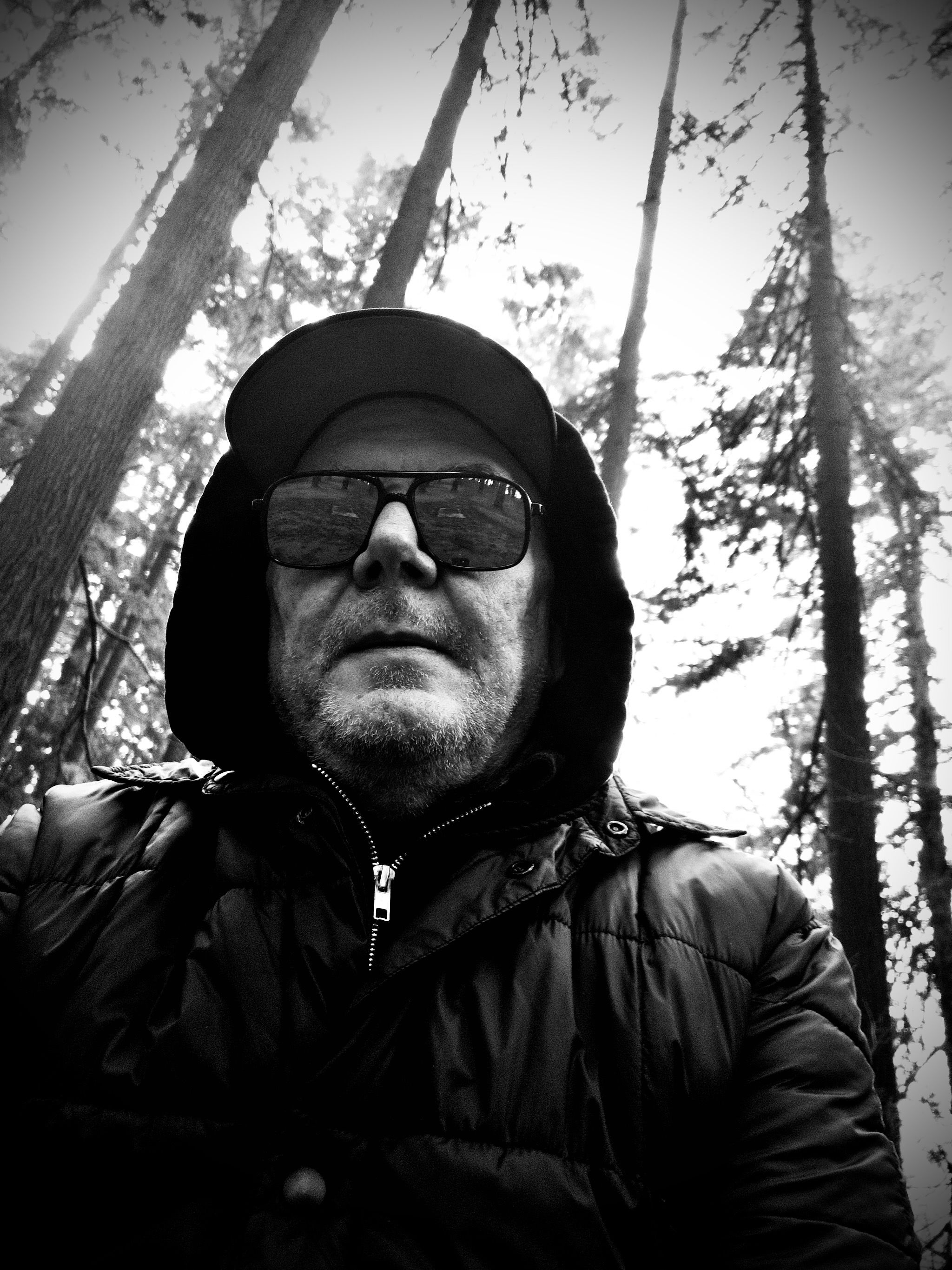 A harsh black and white selfie of me in hoodie, jacket, baseball cap, and sunglasses, with Douglas firs and sky behind me.