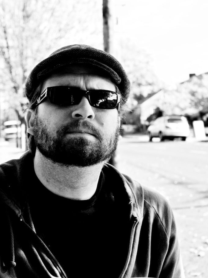In black and white, a white man in hat ans sunglasses, chin slightly up.