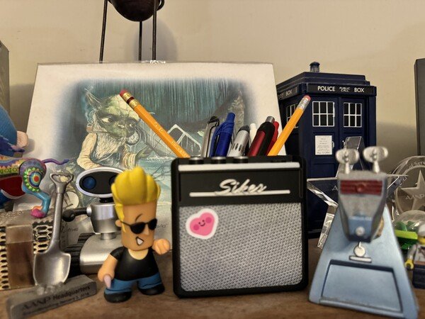 A collection of nerd toys and a jamming speaker with a prami sticker on it. It's being used to hold pens and pencils between Johnny Bravo and K-9.