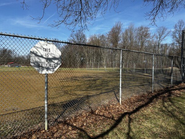 A sign reads 'this is a ballfield'. It is attached to a chainlink fence surrounding a baseball field. 