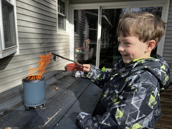 A boy holds a stick over a miniature tabletop fire pit while seated at a blue picnic table.