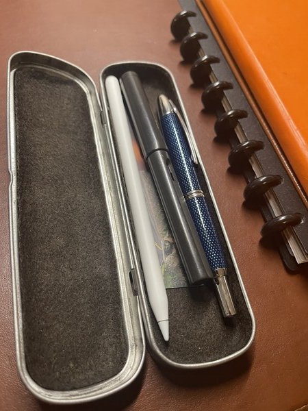 A metal pencil box sitting on a desk next to a stack of notebooks. The box contains an Apple Pencil and two fountain pens.