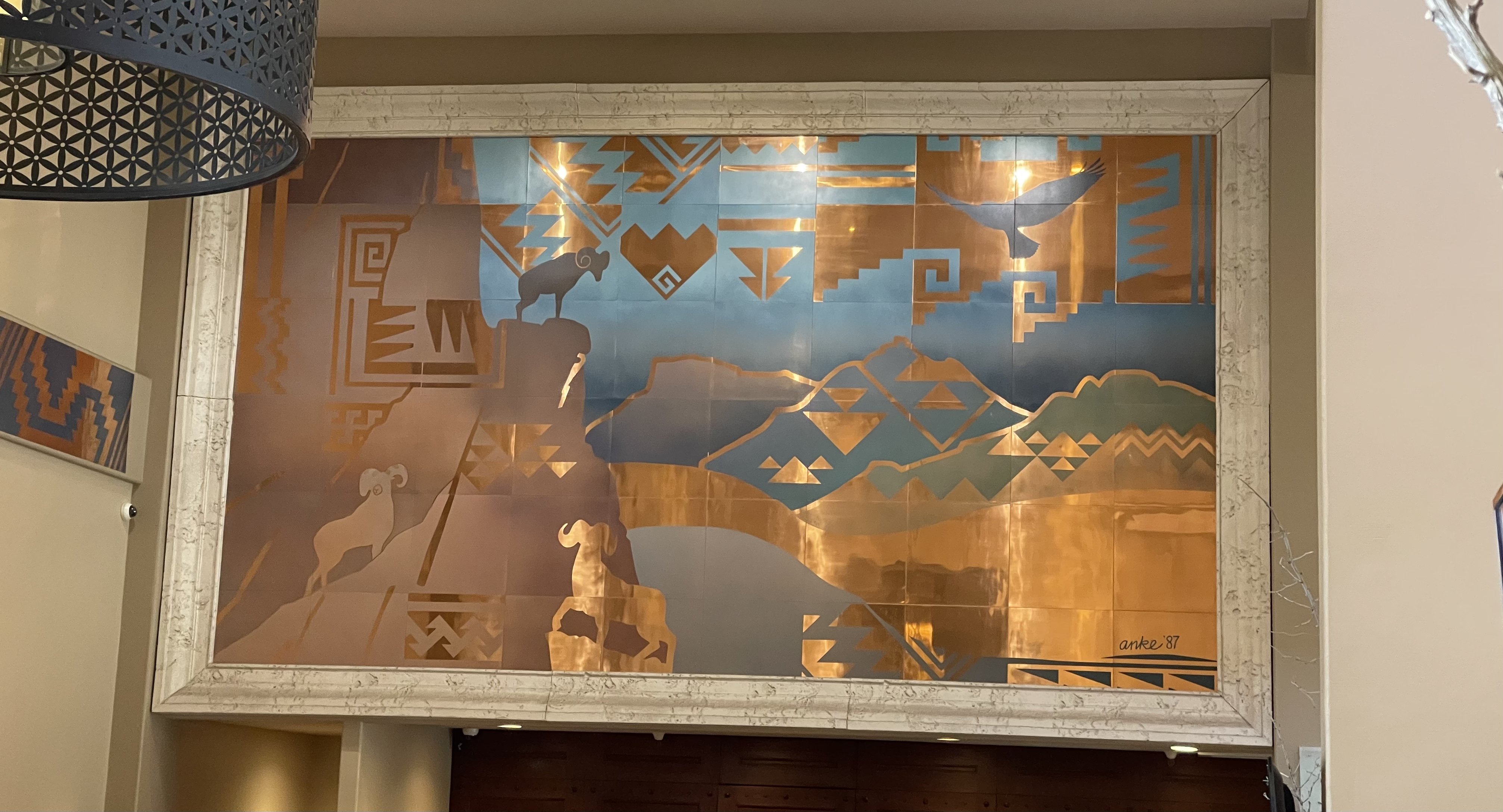 A large mural in copper and turquoise, high on a wall, depicting a Southwest scene with bighorn sheep. 