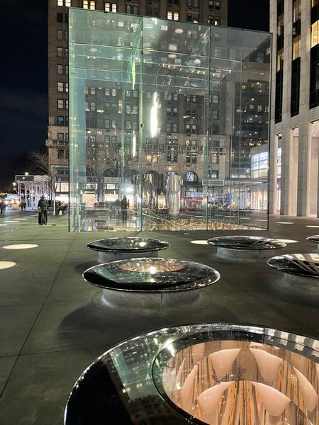 A night time photo of the Apple Store glass cube on 5th Avenue in Manhattan. Glass skylight features appear in the foreground. 