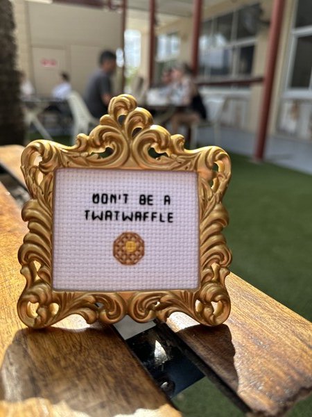 A small picture frame with ornate gold leafing around the frame itself sits atop a table.  It’s about 3” x 3”, and it has a small cross stitch waffle along with the words “Don’t be a twatwaffle” stitched into it.
