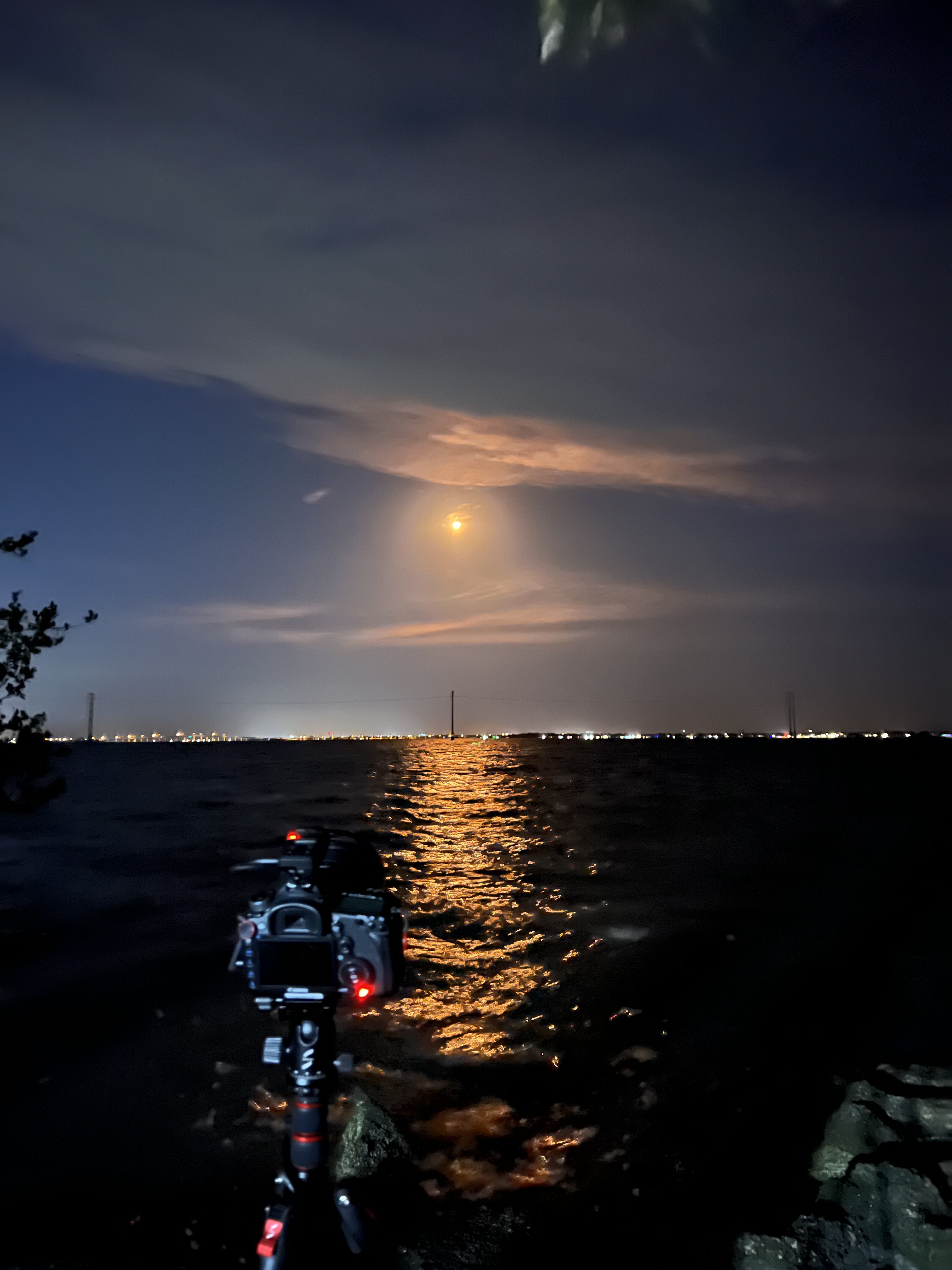 SpaceX rocket launch at night, the orange glow of the rocket plume reflecting on the water from the river in the foreground. A black digital camera on a tripod in the foreground captures the event.