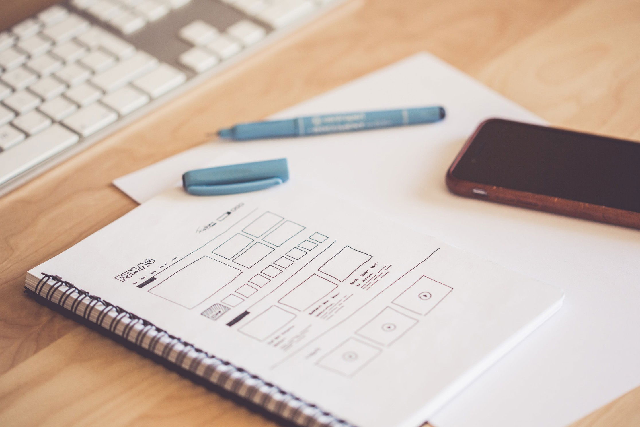 A notebook on a wooden desk with a keyboard pen and phone slightly out-of-focus. The notebook is showing a hand-drawn sketch of a website titled "FJMAG" with many boxes presumably representing images and articles, as well as ones with "play" buttons representing videos.