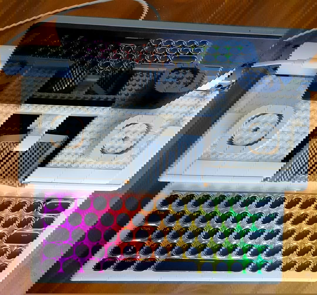 A top-down shot of a white, Lego-built typewriter with an RGB mechanical keyboard for the keys. The keys have rounded keycaps. There is a tablet sitting on top as if it was the paper and is plugged in via USB. The keys and other reflections are reflected in the blank screen