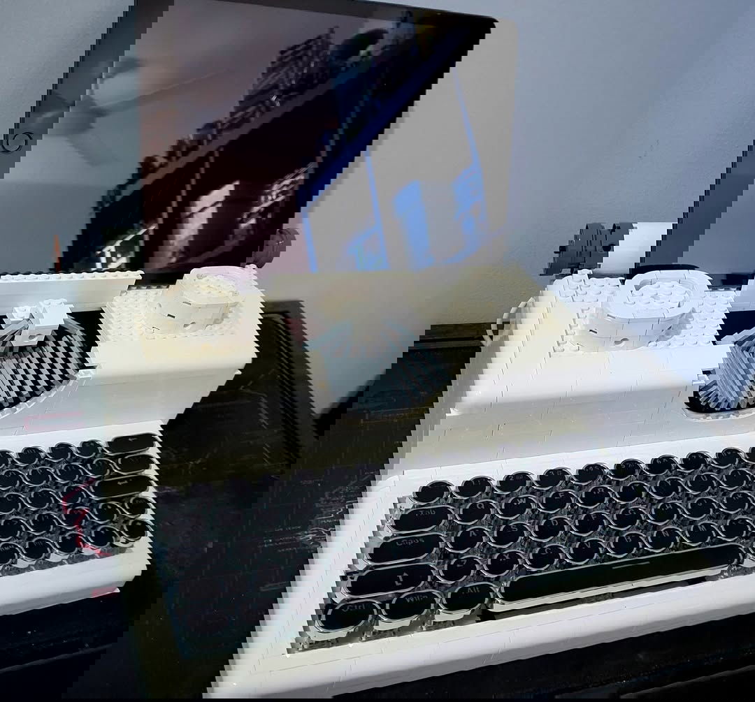 A front-on shot of a white, Lego-built typewriter with an RGB mechanical keyboard for the keys. The keys have rounded keycaps. There is a 10" iPad sitting on top as if it was the paper. It's on a desk against a white wall.