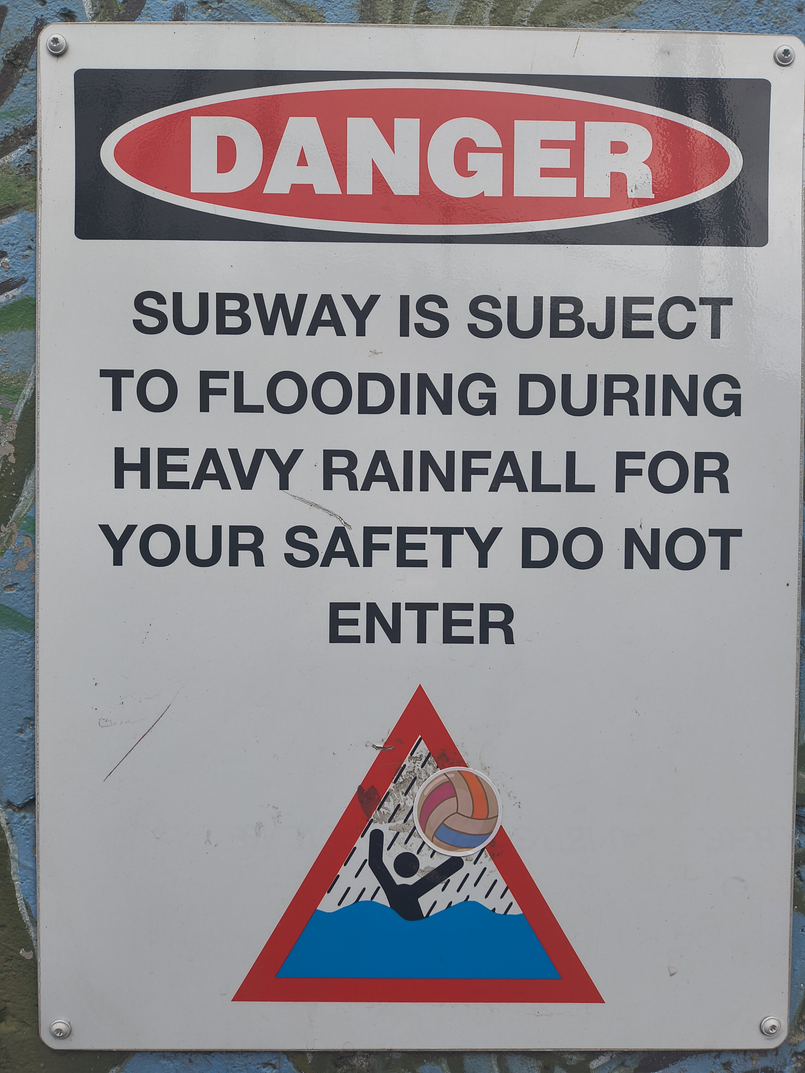 A flood warning sign has a ball sticker, making it look like the drowning figure is instead playing water polo 😆