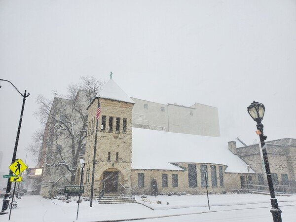 Through blowing snow, a stone church with a gray boxy building looming behind, snow snow snow. 