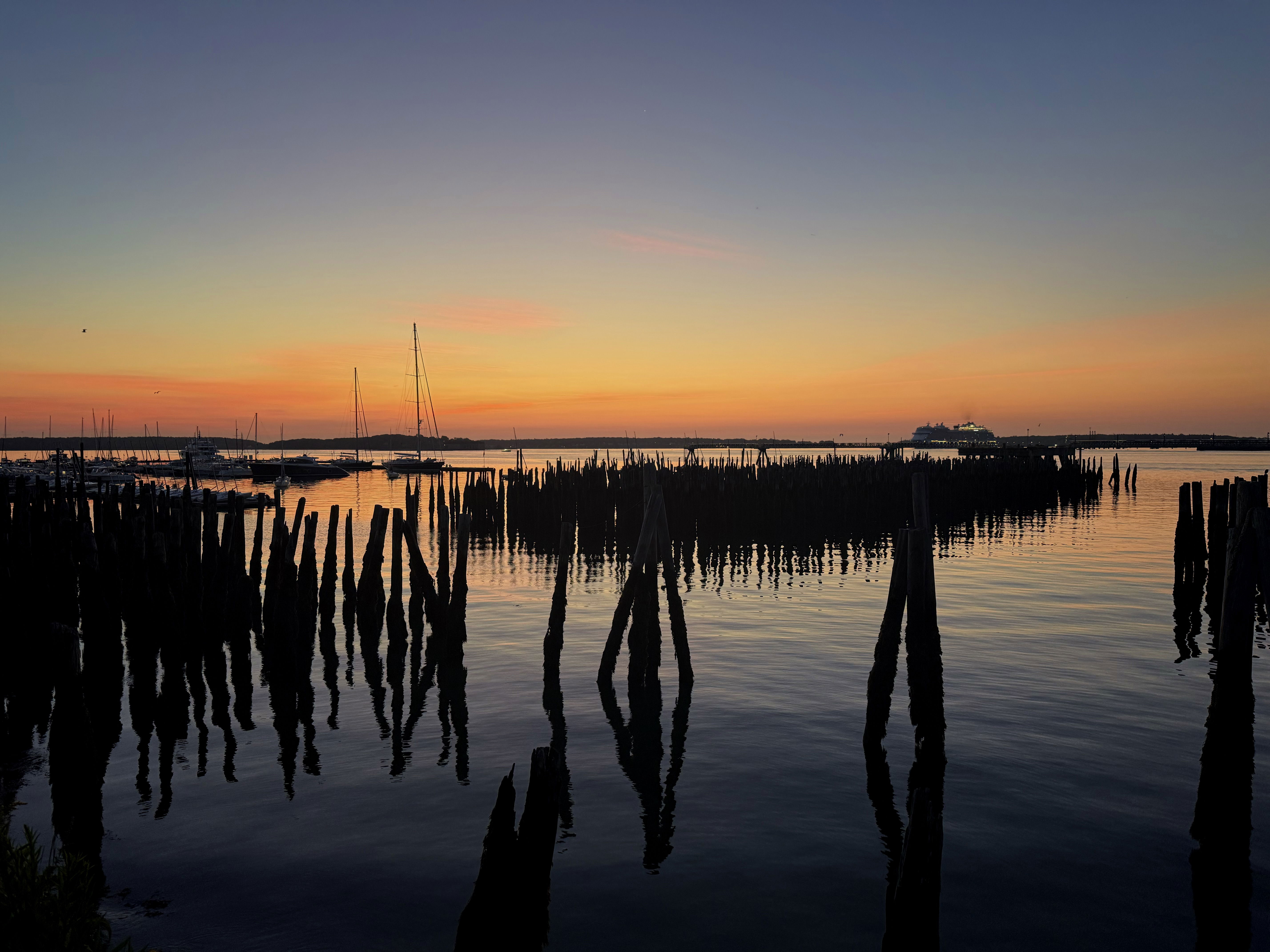 Silhouette of old wooden pilings in the foreground, reflecting in still water. A colorful sunset with shades of orange and purple in the sky, alongside distant boats in the harbor.
