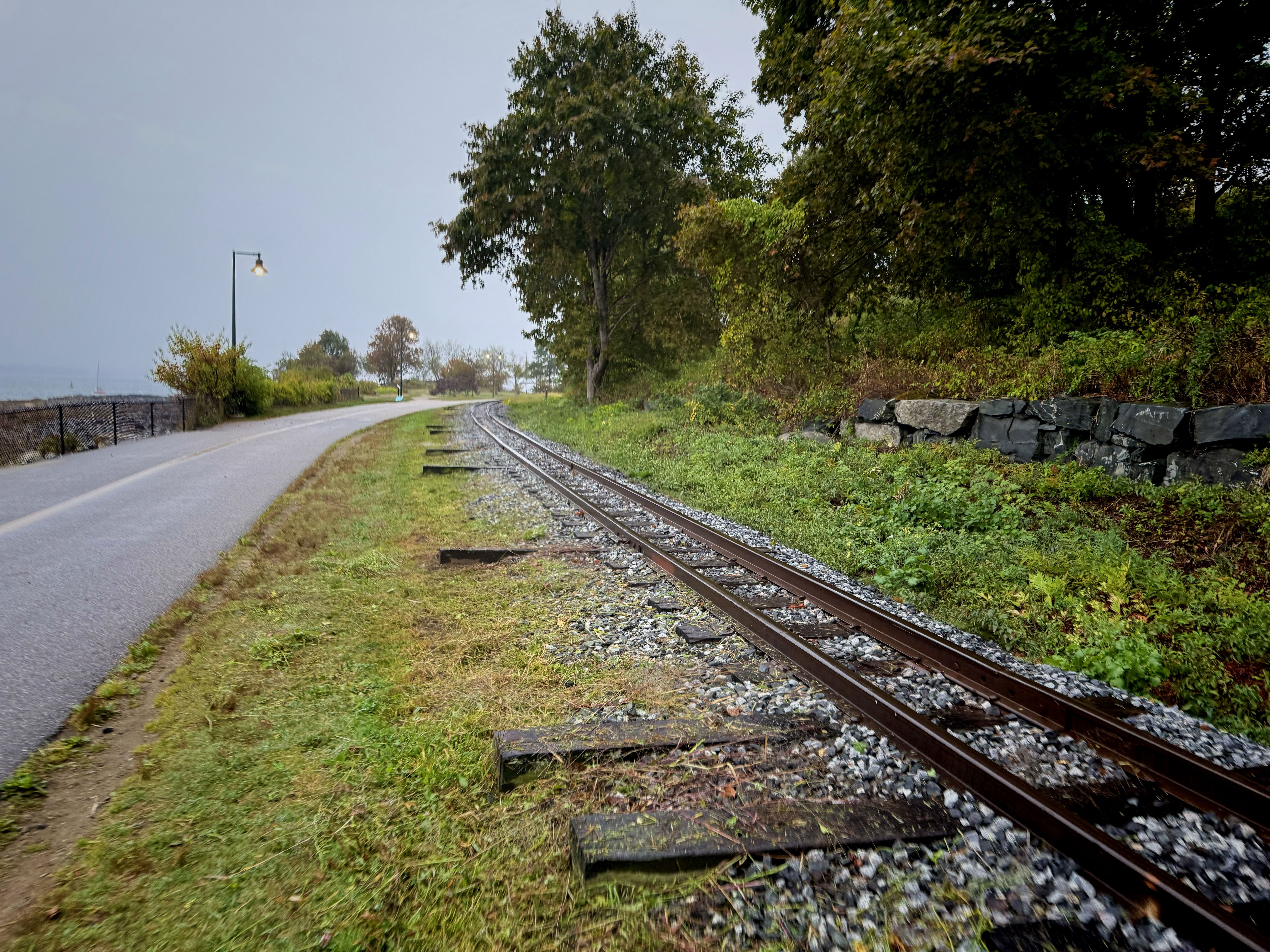 A view of a gravel path alongside a set of railway tracks, with trees and greenery on one side and a fence along a body of water in the distance. A light pole is visible along the path, and the sky appears overcast.