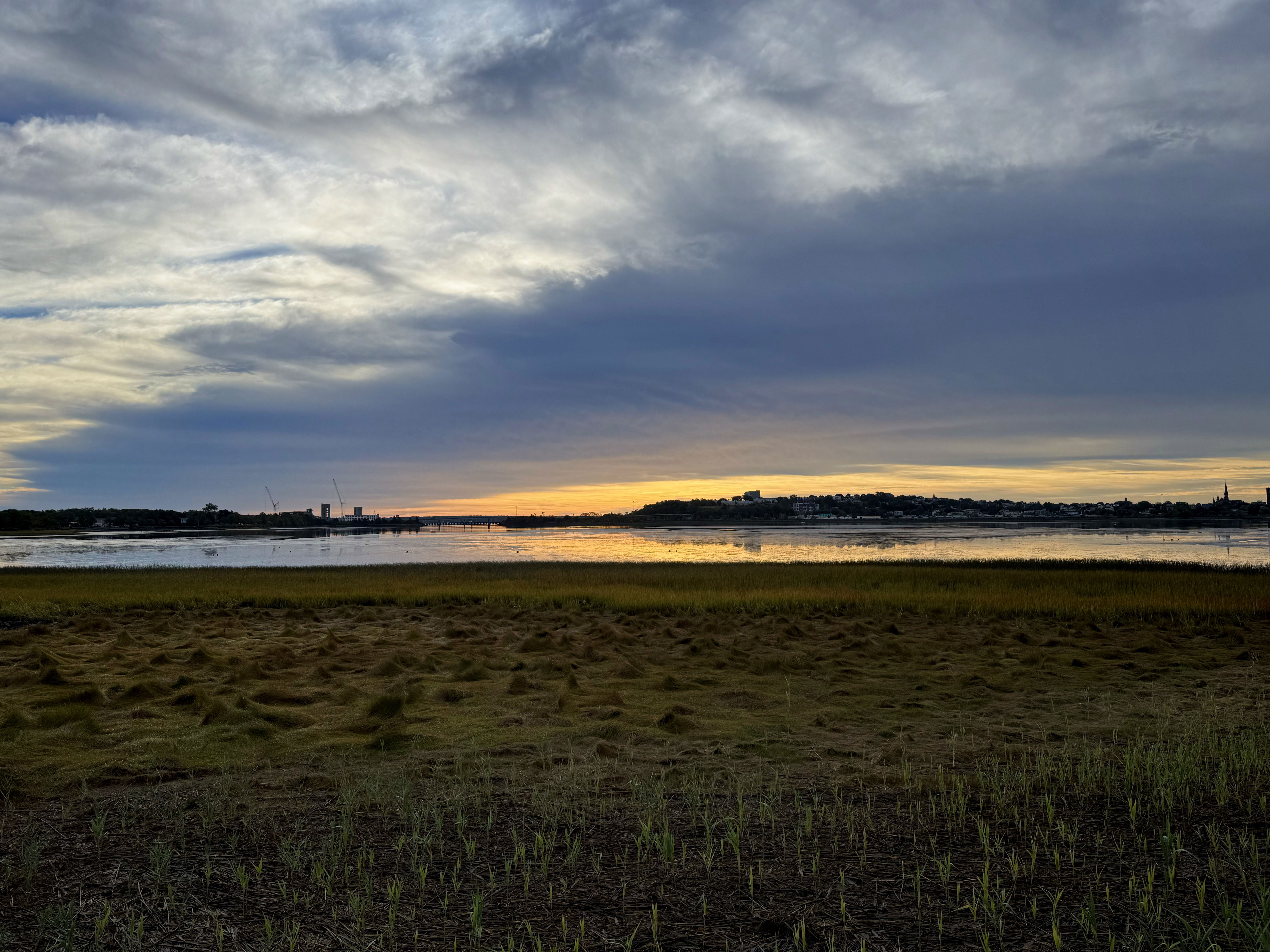 A scenic view of a calm river at sunset, with dark clouds above and a soft orange glow on the horizon. The foreground features grassy land leading to the water, while the background shows a silhouette of buildings and trees.