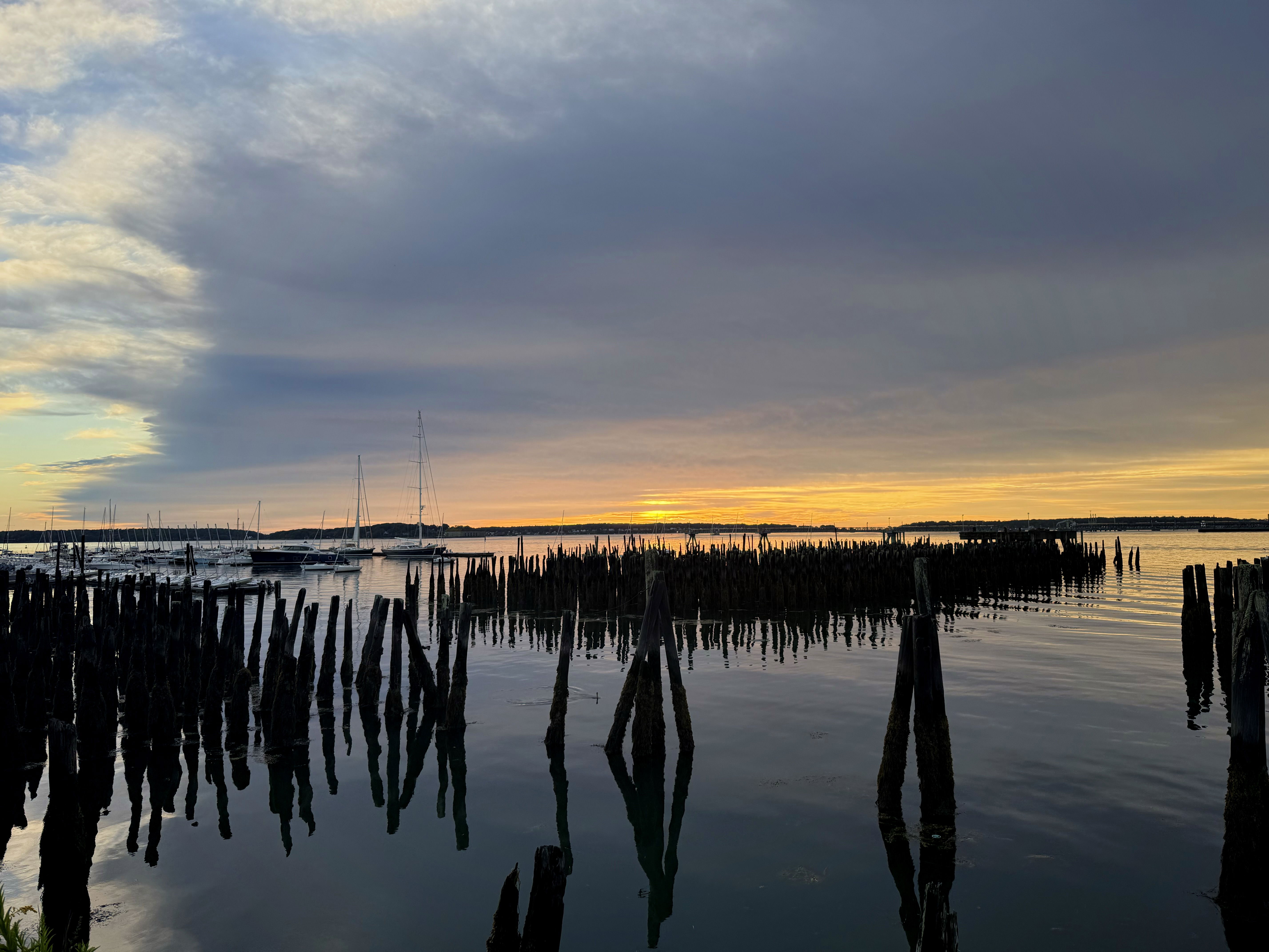 A tranquil sunset over a calm body of water, featuring old wooden pilings sticking out from the water’s surface. Sailboats are moored in the background against a colorful sky filled with clouds.