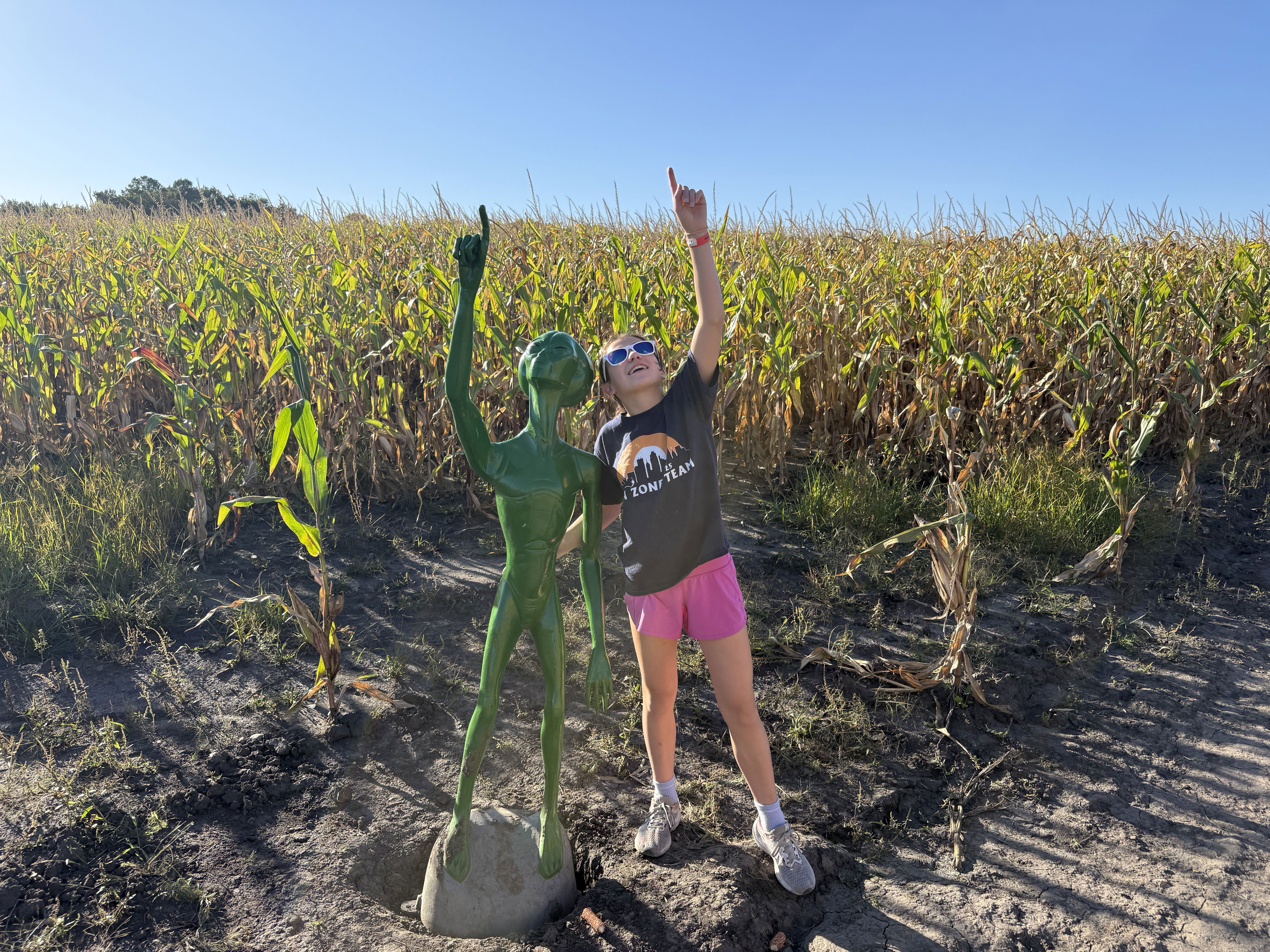 A young girl stands beside a green alien statue in a cornfield, both pointing upwards towards the sky. The sun is shining and the corn plants are visible in the background.