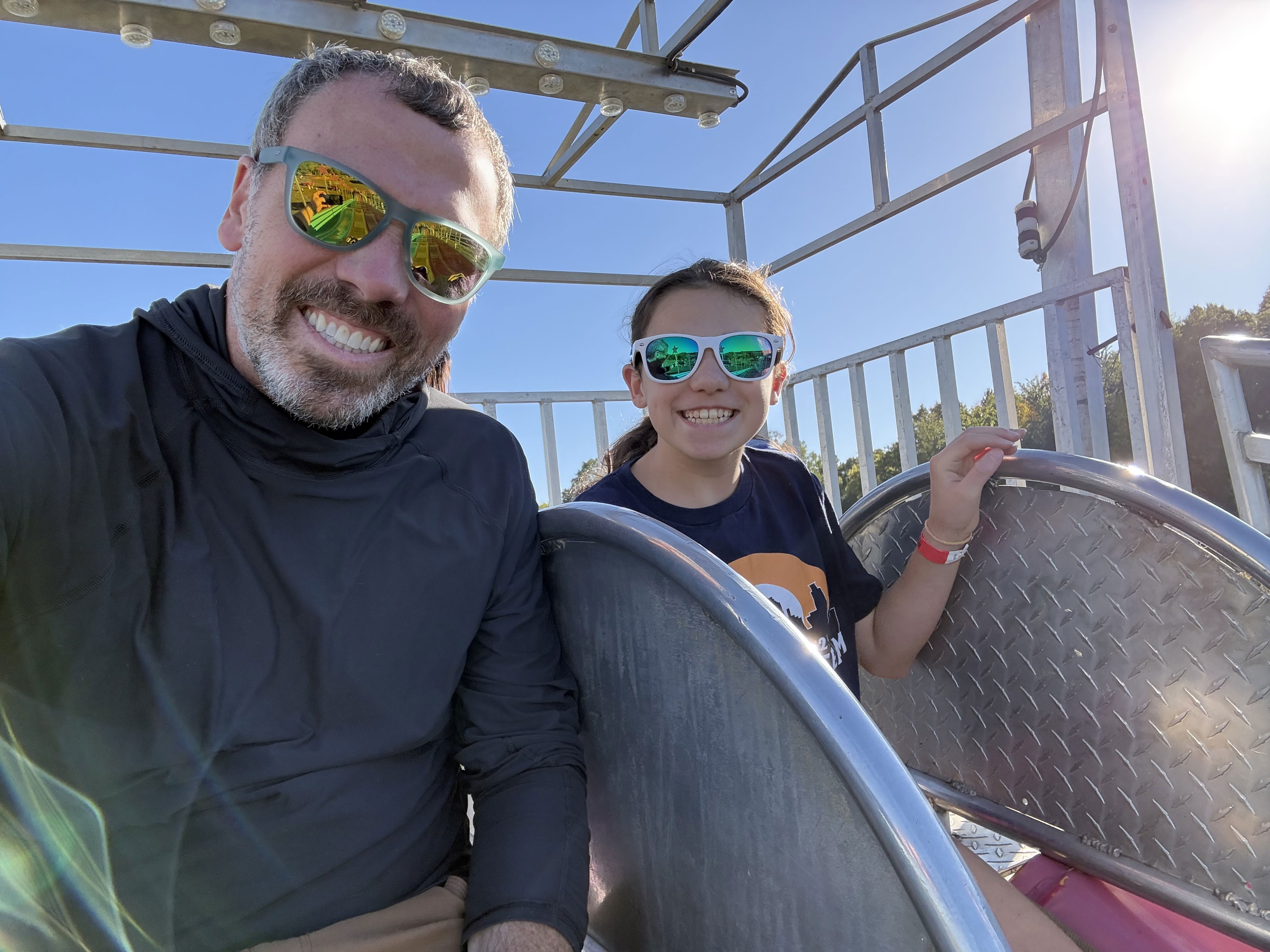 A smiling man and girl wearing sunglasses sit side by side in a metal ride structure on a sunny day. The background features a bright blue sky and hints of greenery.