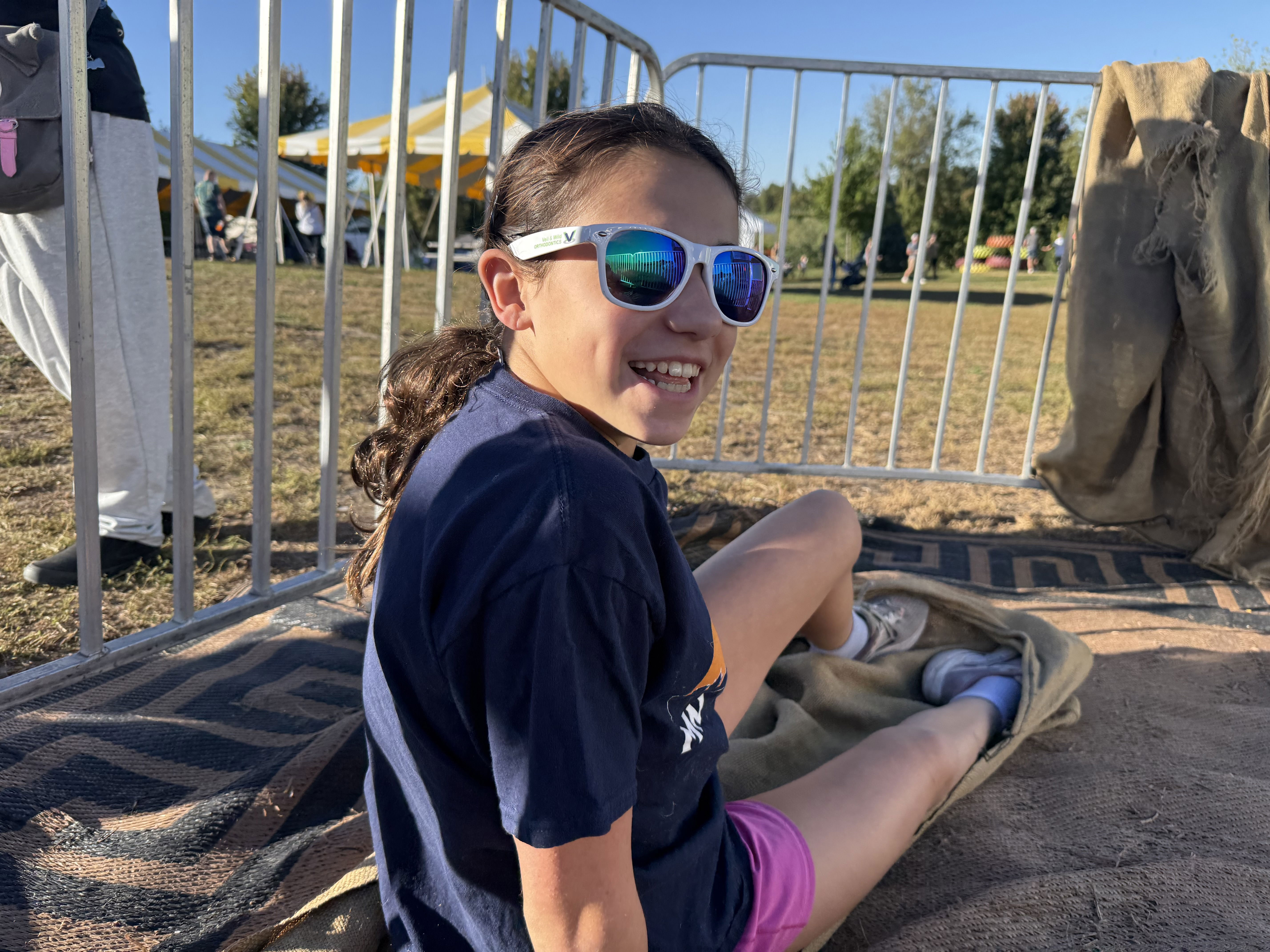 A young girl with long hair sits on the grass, wearing sunglasses and a navy shirt. She is smiling and appears relaxed, with a blanket partially covering her legs. In the background, there are people and a yellow tent.