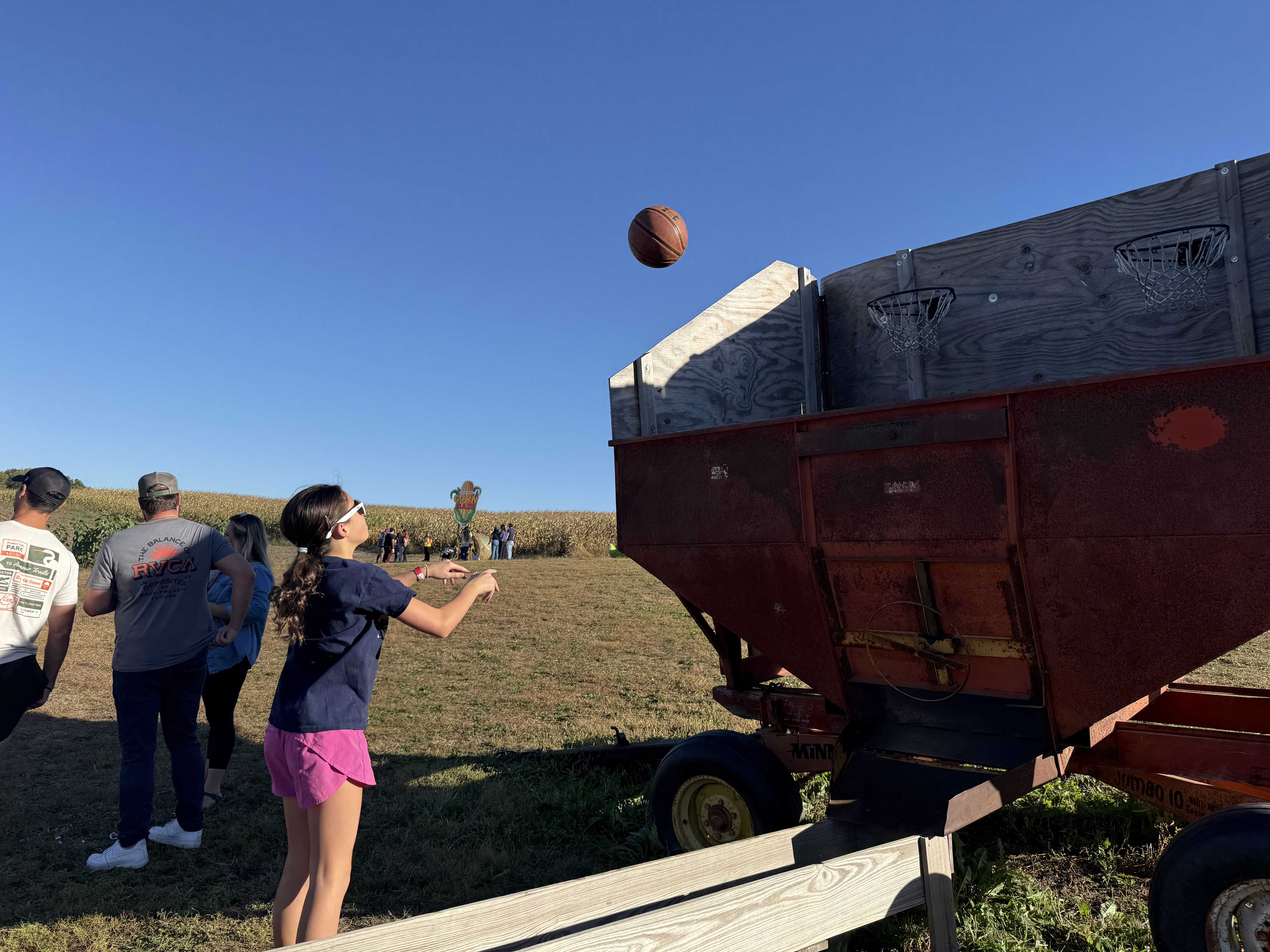 A girl in a navy shirt and pink shorts is shooting a basketball towards a farm equipment with basketball hoops attached. In the background, a few people are watching, surrounded by corn fields under a clear blue sky.
