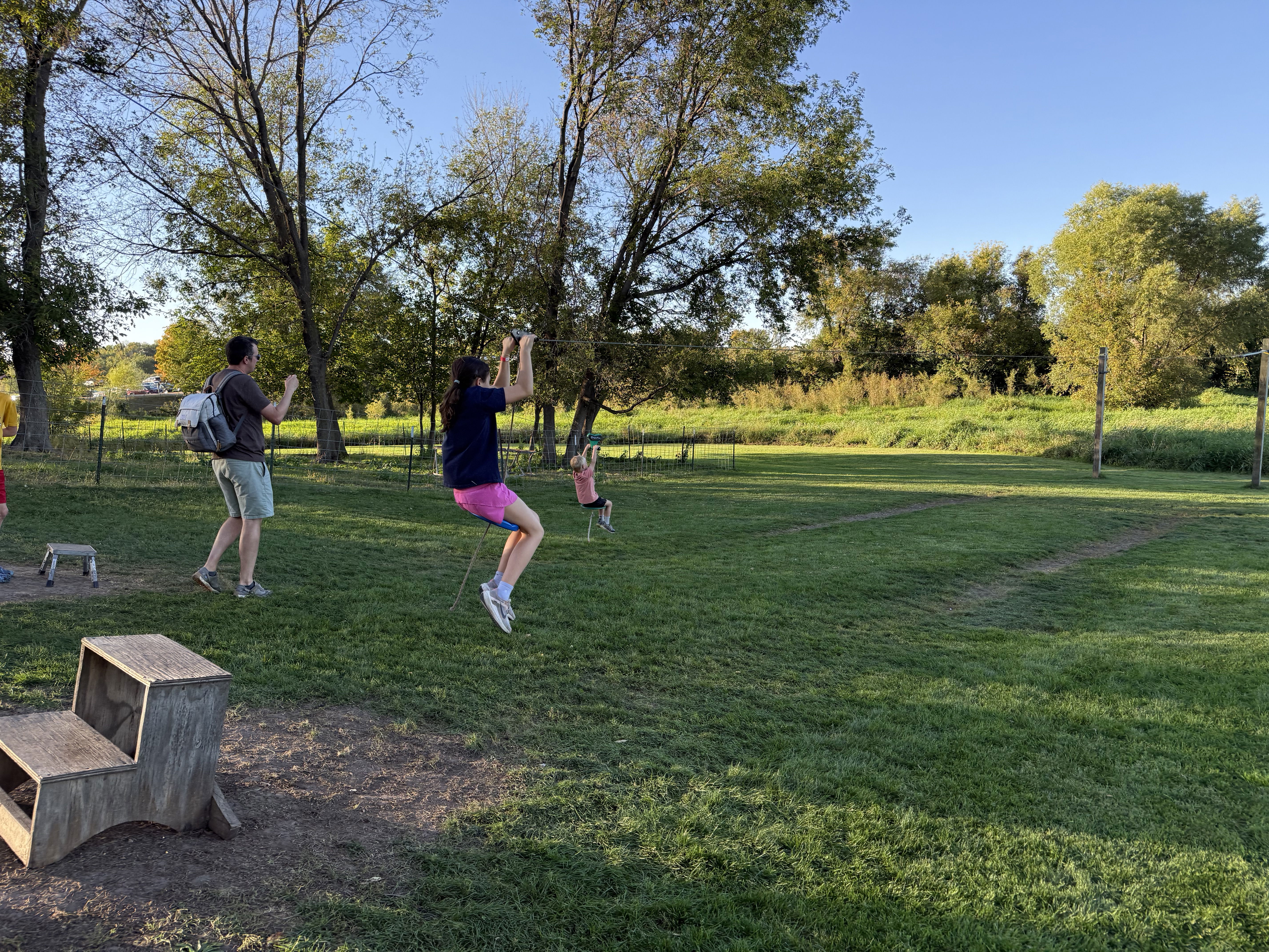 A group of children and an adult enjoy a grassy outdoor area, with one child on a zip line and another on a nearby swing. Trees line the background under a clear blue sky.