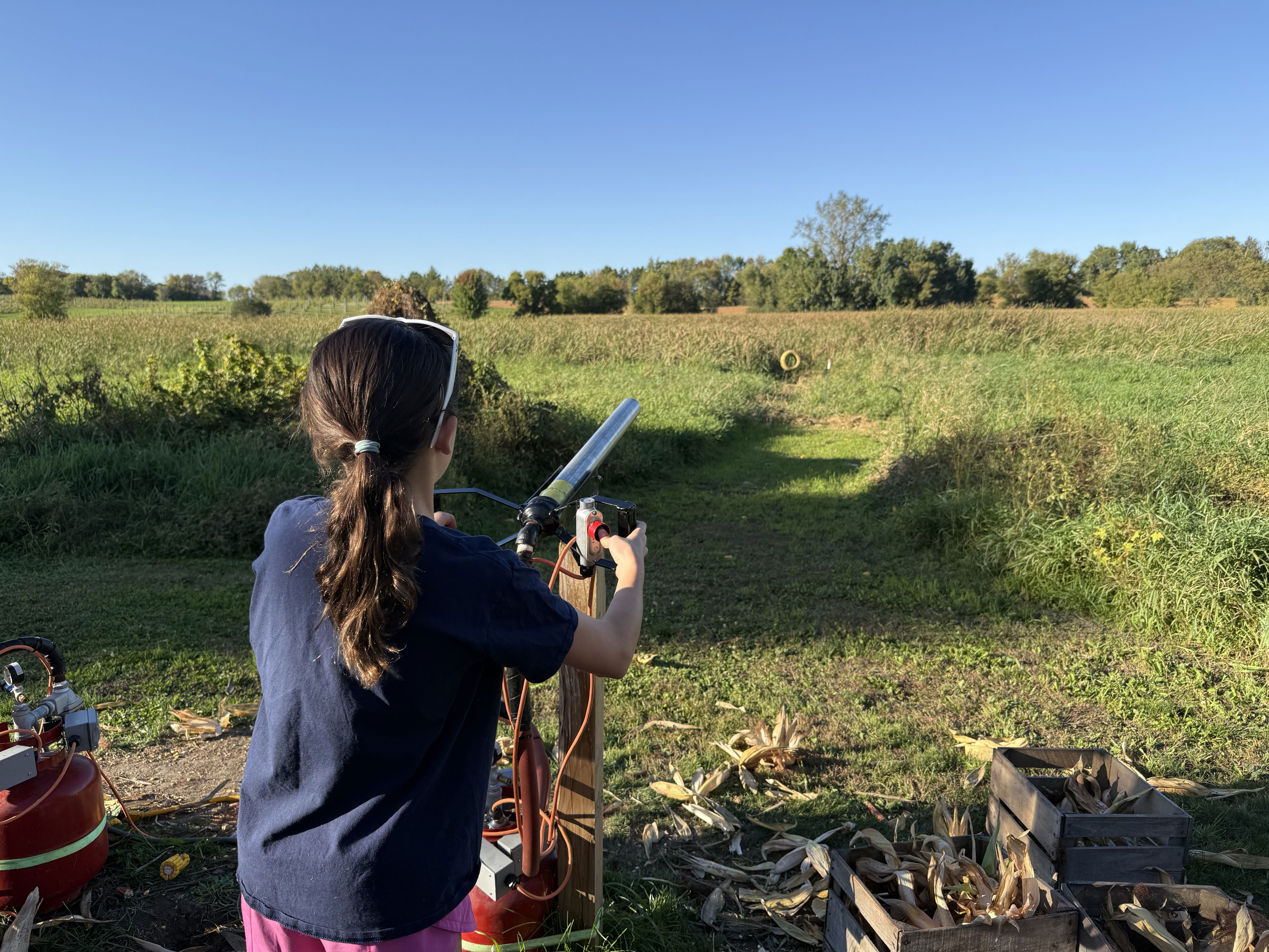 A young girl stands with her back to the camera, aiming a launcher at a distant target in an open grassy field surrounded by trees. The sky is clear and sunny, and there are equipment and corn husks nearby.