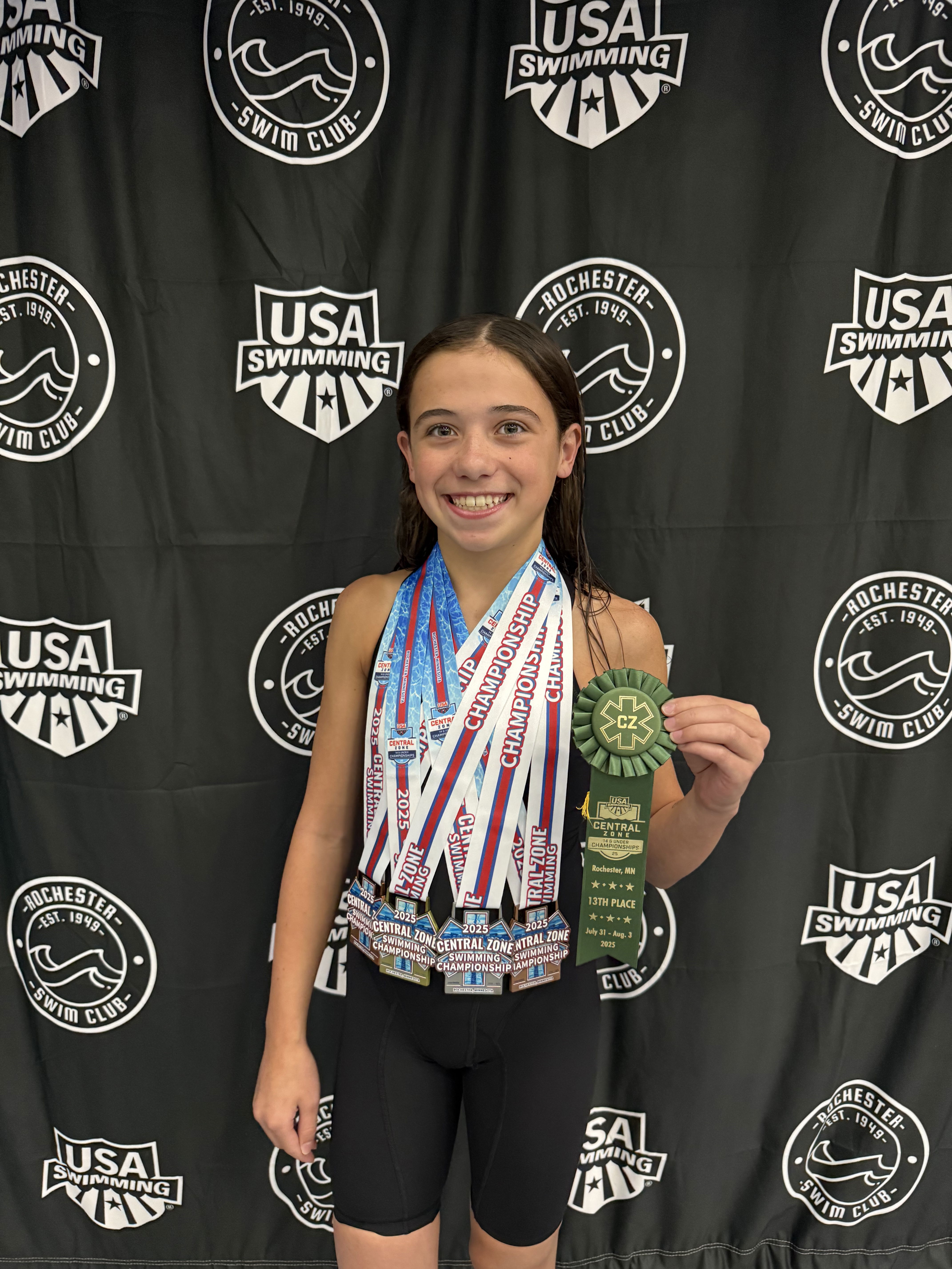 A young swimmer stands smiling with numerous medals draped around her neck and a green ribbon for 13th place in her hand. She is in front of a background featuring logos of USA Swimming and the Rochester Swim Club.