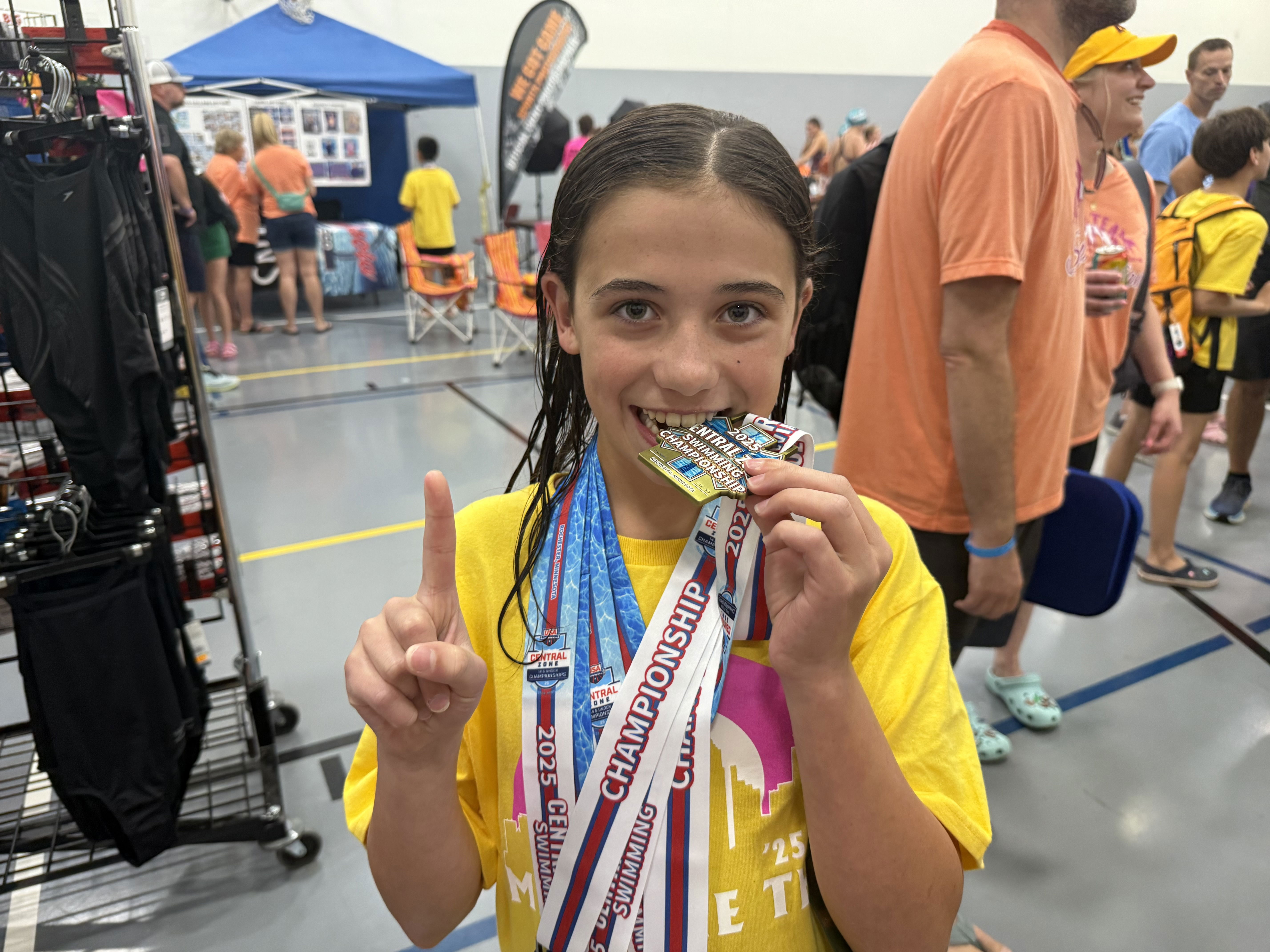 A young girl wearing a bright yellow t-shirt smiles while holding a medal in her mouth and showing a “number one” sign with her finger. She is adorned with multiple medals draped around her neck, standing in a busy indoor setting with people and