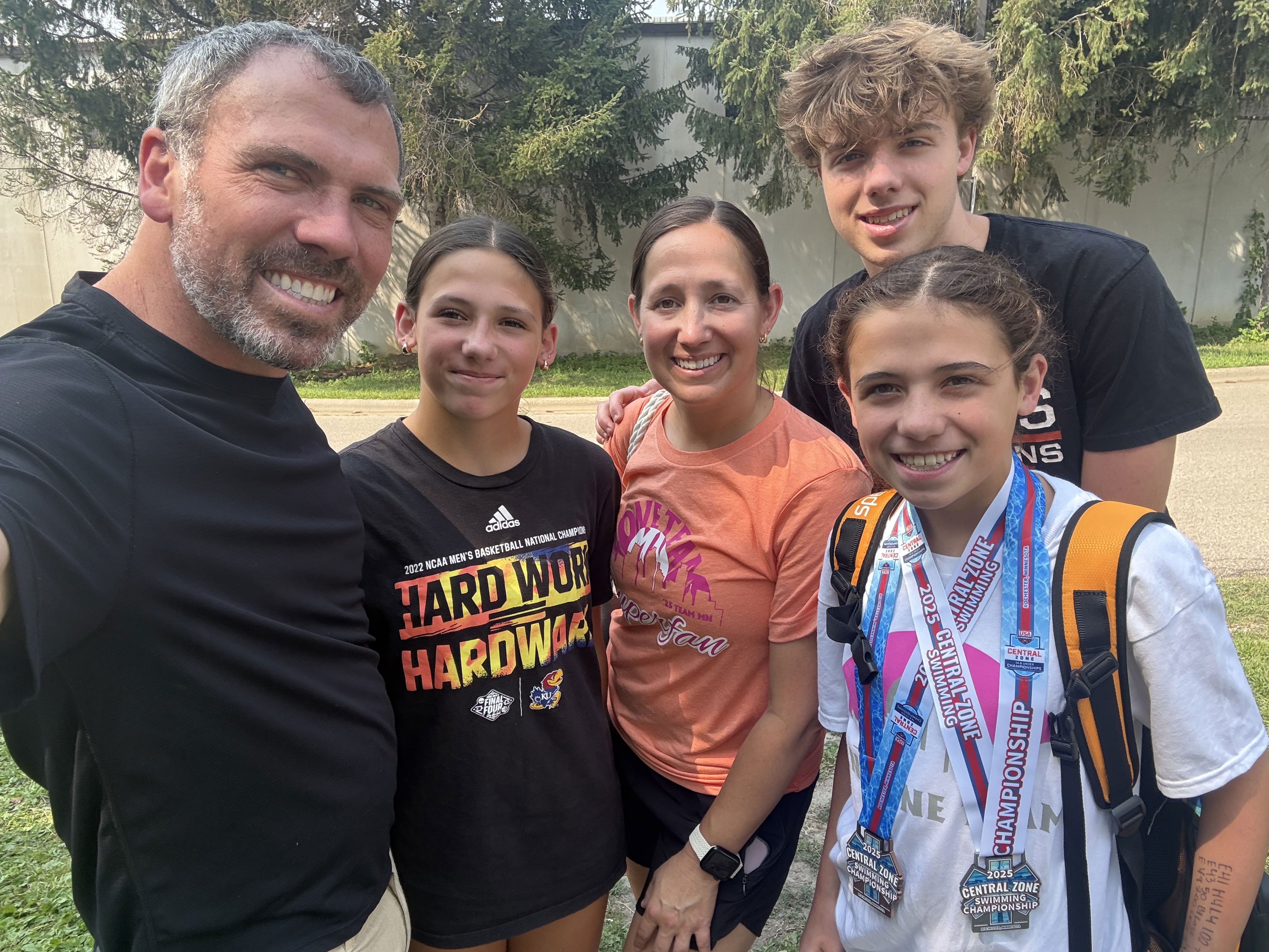 A group photo featuring five individuals smiling together outdoors. They display a sense of celebration, with one girl wearing multiple medals around her neck. The group includes three girls and two adults, with trees in the background.