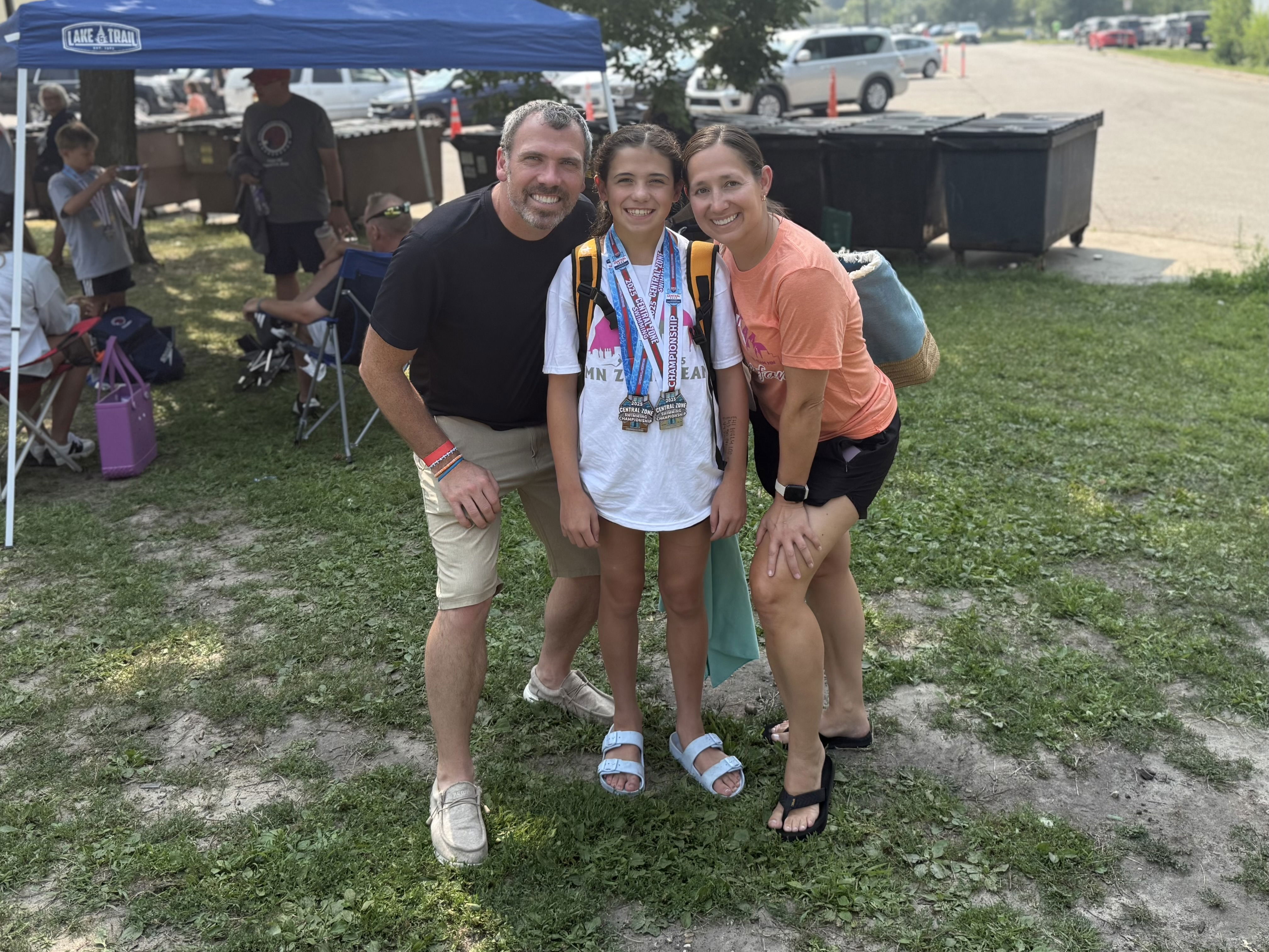 A young girl stands proudly in the center, wearing multiple medals around her neck, flanked by two adults who are smiling at the camera. The background features a blue tent, grass, and a few people sitting in chairs.
