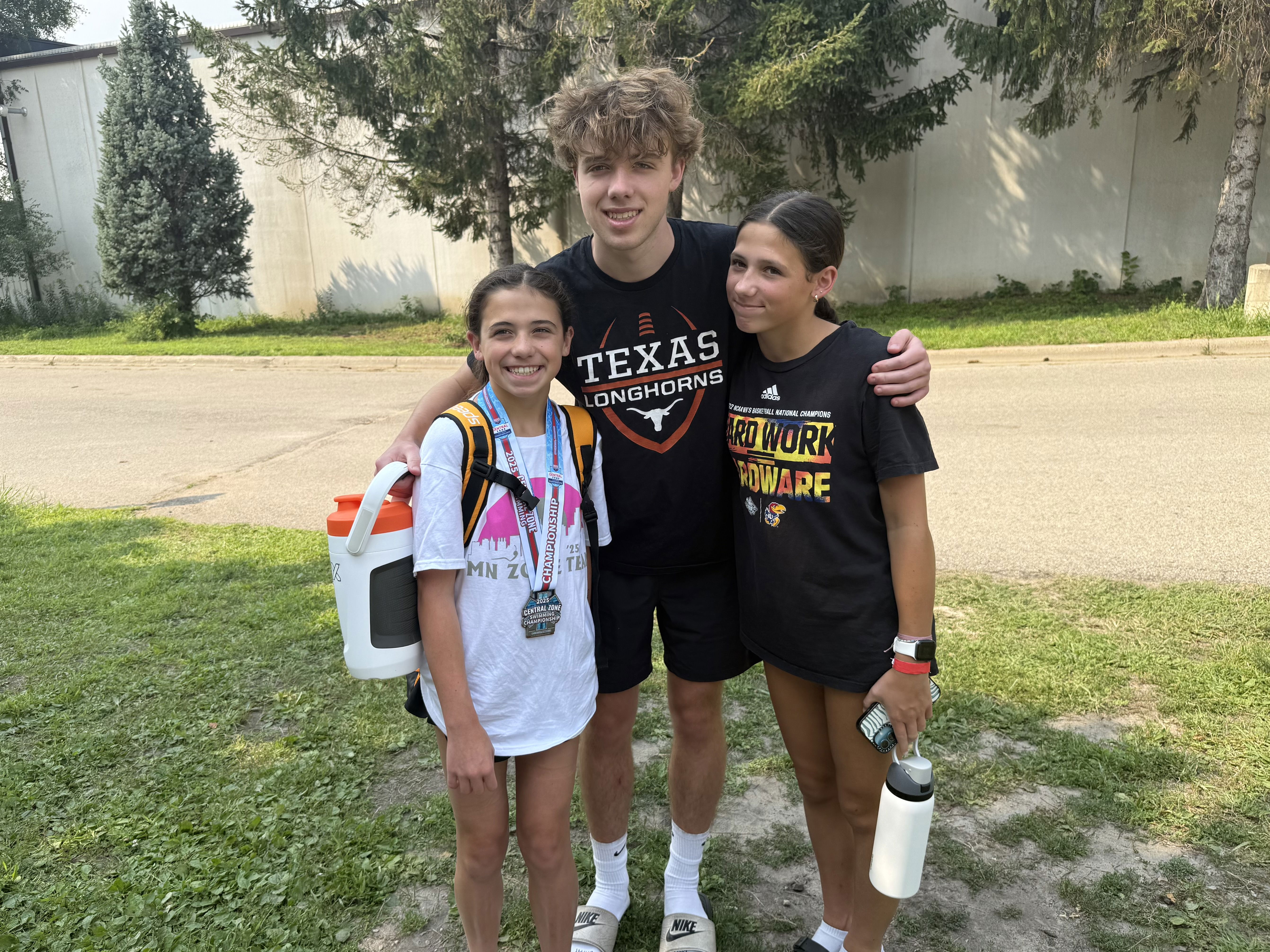 Three young people pose together outdoors. Two girls are smiling, one wearing a medal and a backpack, and the other holding a water bottle. A boy in a black t-shirt stands in the middle with his arm around one girl. They are in a