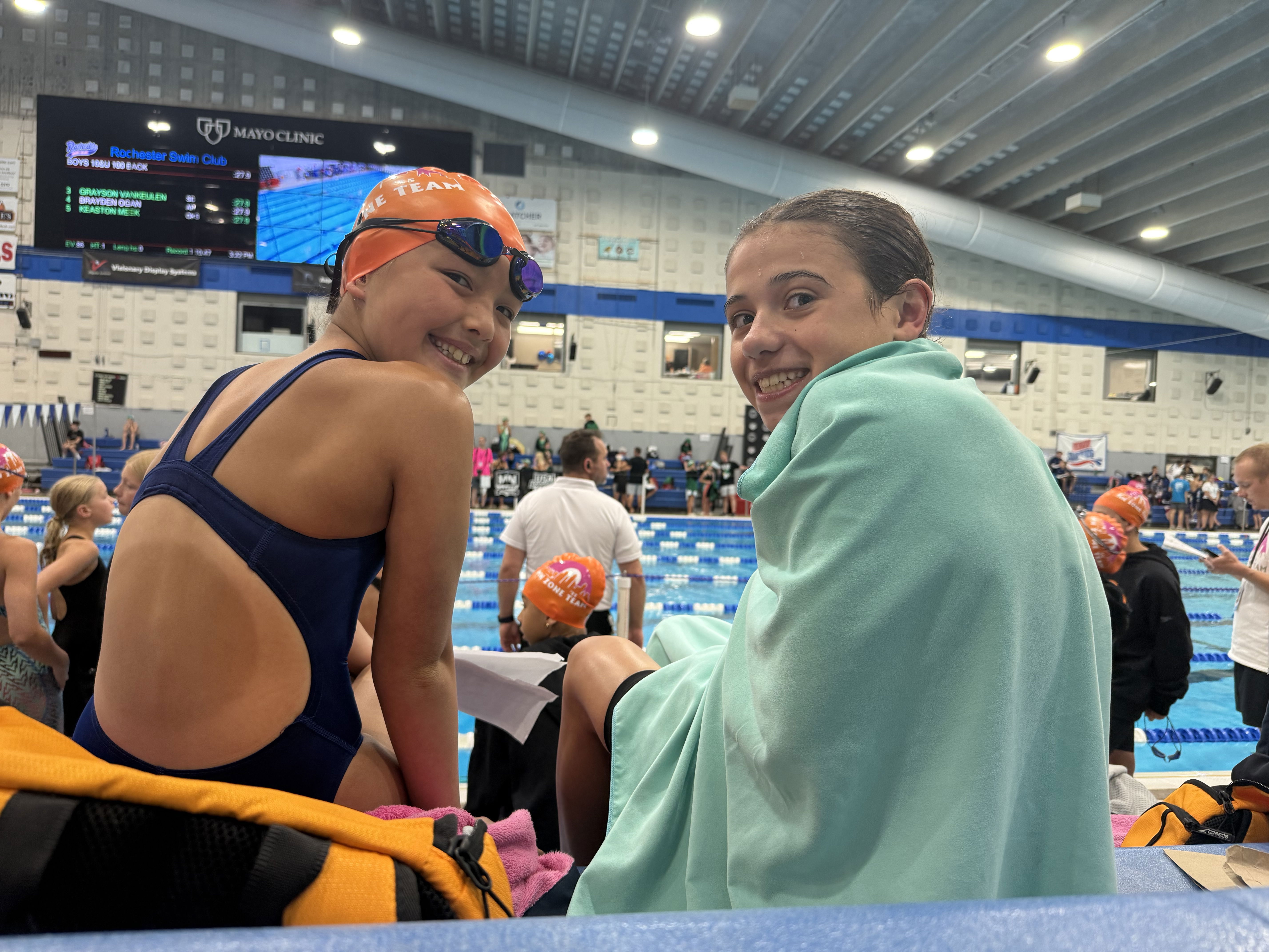 Two young swimmers sit together at a poolside during a competition. One girl, wearing a swim cap and goggles, smiles brightly over her shoulder, while the other, wrapped in a light blue towel, grins at the camera. The background features a