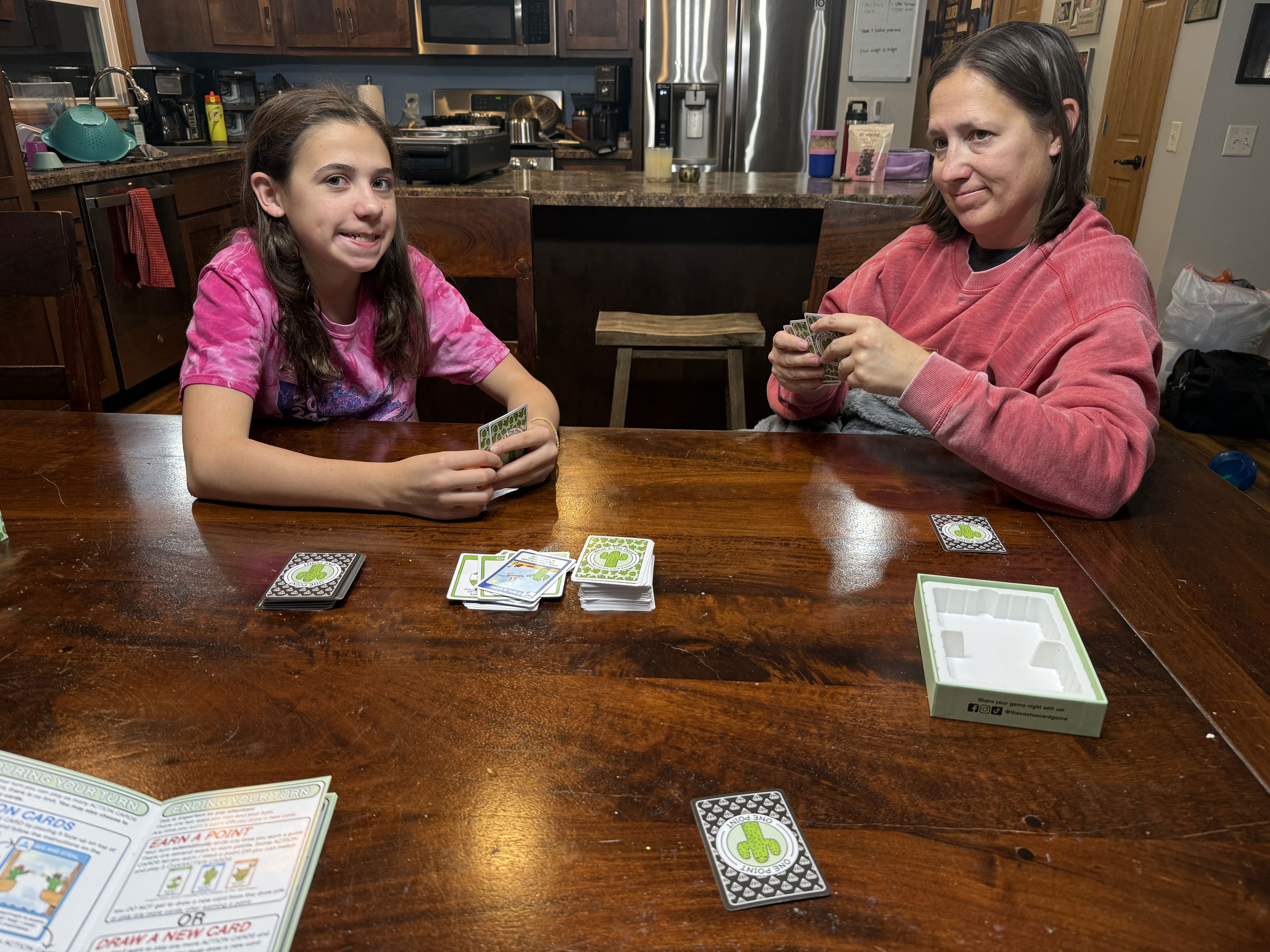 A girl and a woman are seated at a wooden table, playing a card game. The girl, wearing a pink tie-dye shirt, is smiling and holding cards, while the woman in a red sweatshirt looks at her cards with a thoughtful expression.
