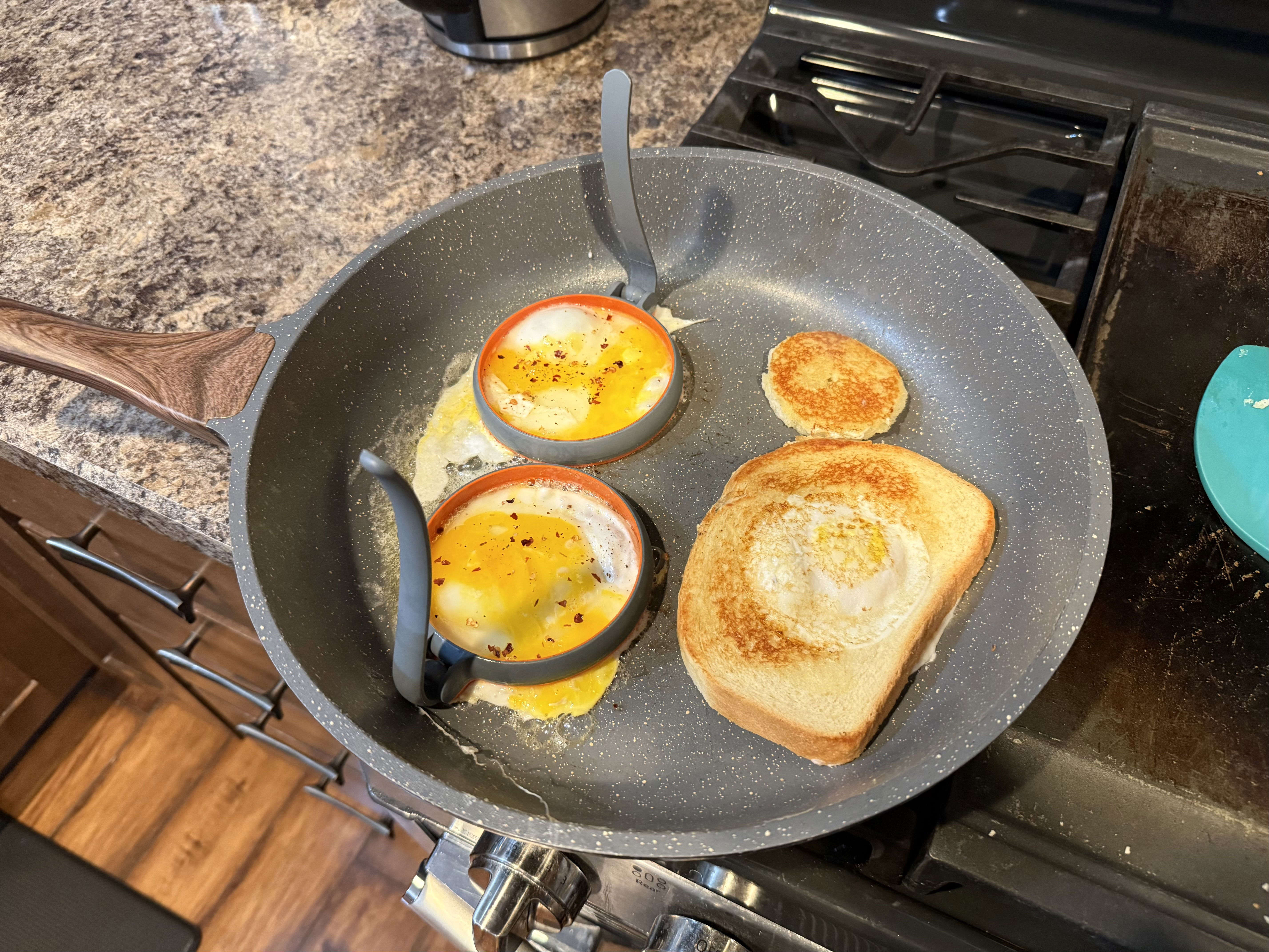 Frying pan on a stove with two eggs in round molds, a piece of toasted bread, and a small round piece of toasted bread, all on a granite countertop.