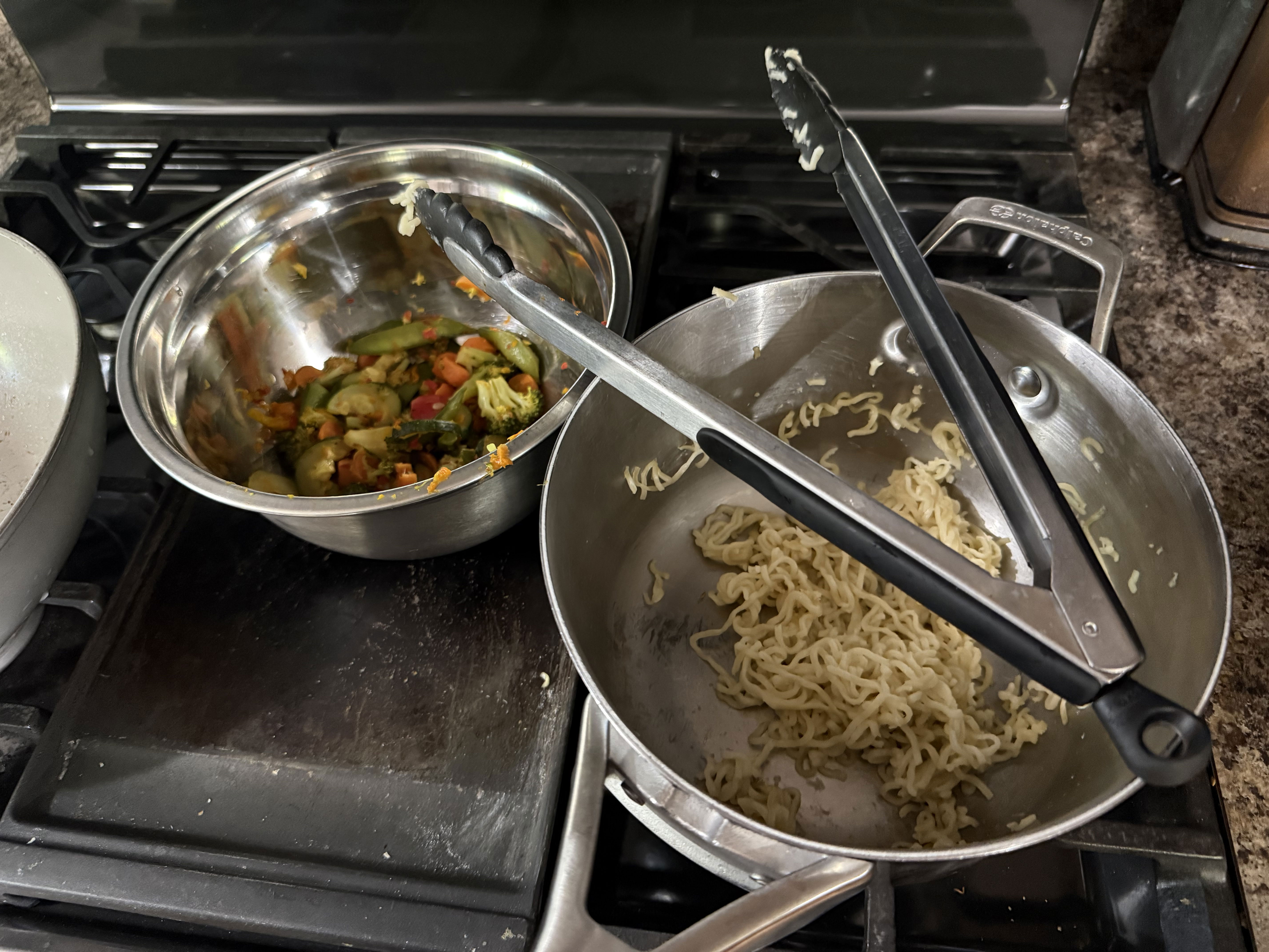 A stove with two pots: one containing cooked noodles and the other with mixed vegetables. Both pots have tongs resting on them.