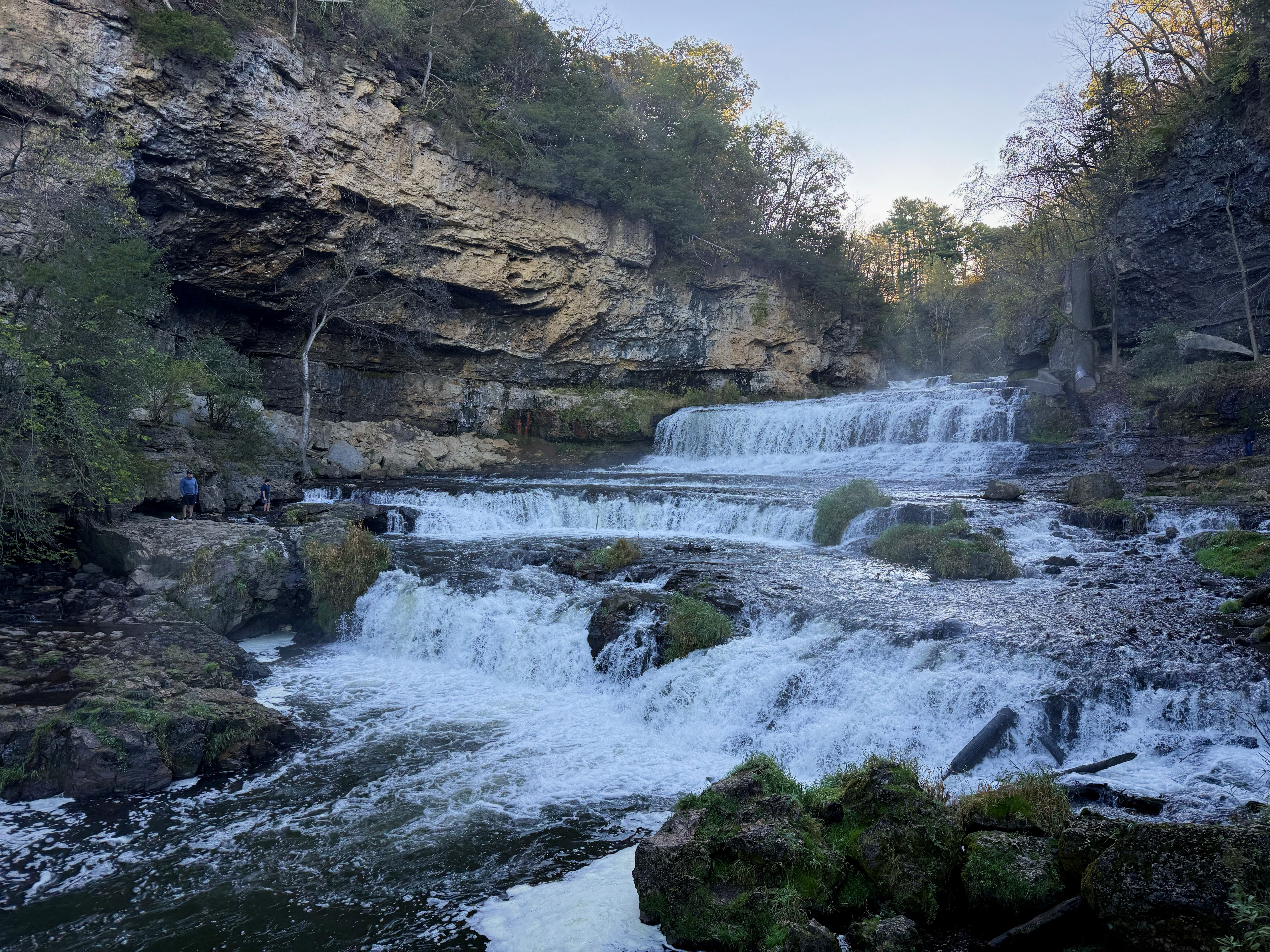 A scenic view of cascading waterfalls surrounded by rocky cliffs and greenery, with two people walking along a path near the water’s edge. The sky is clear, indicating a peaceful outdoor setting.