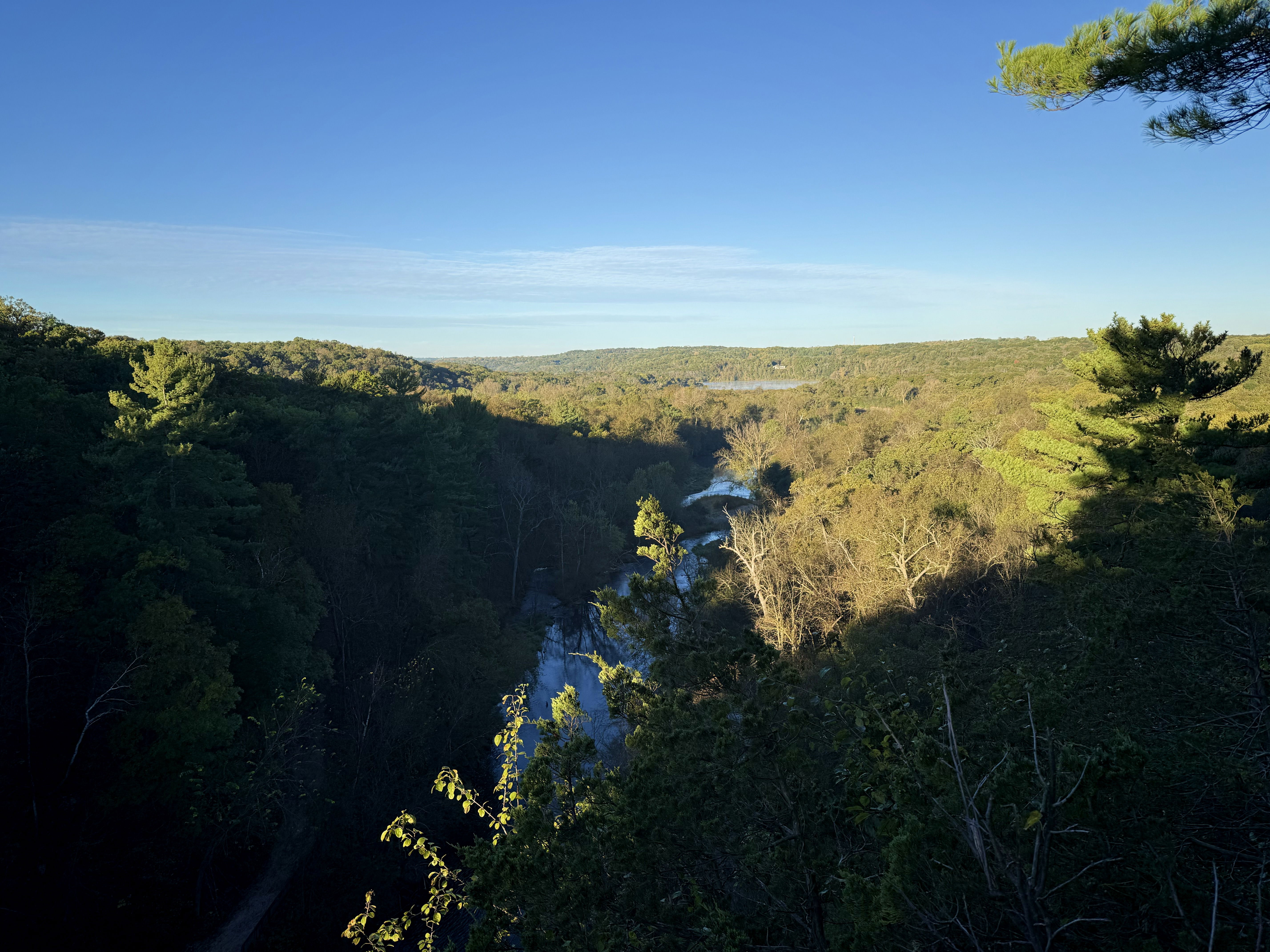 A scenic view of a river winding through a dense forest, with hills in the background under a clear blue sky. The landscape features a mix of green pine and autumn-colored trees. Shadows from the trees add depth to the image.